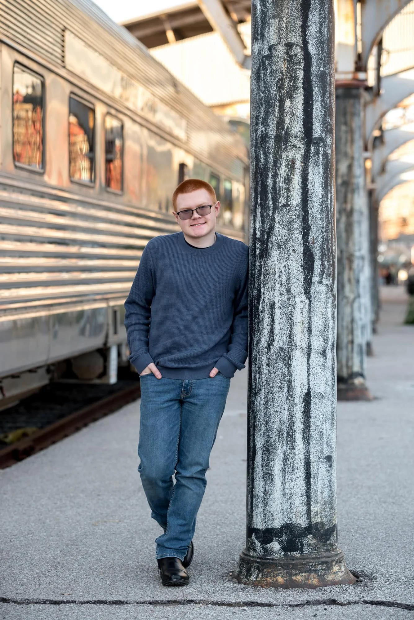 Senior picture taken by a St. Louis City photographer showing a boy in a blue shirt leaning against a pole with a train car behind him.