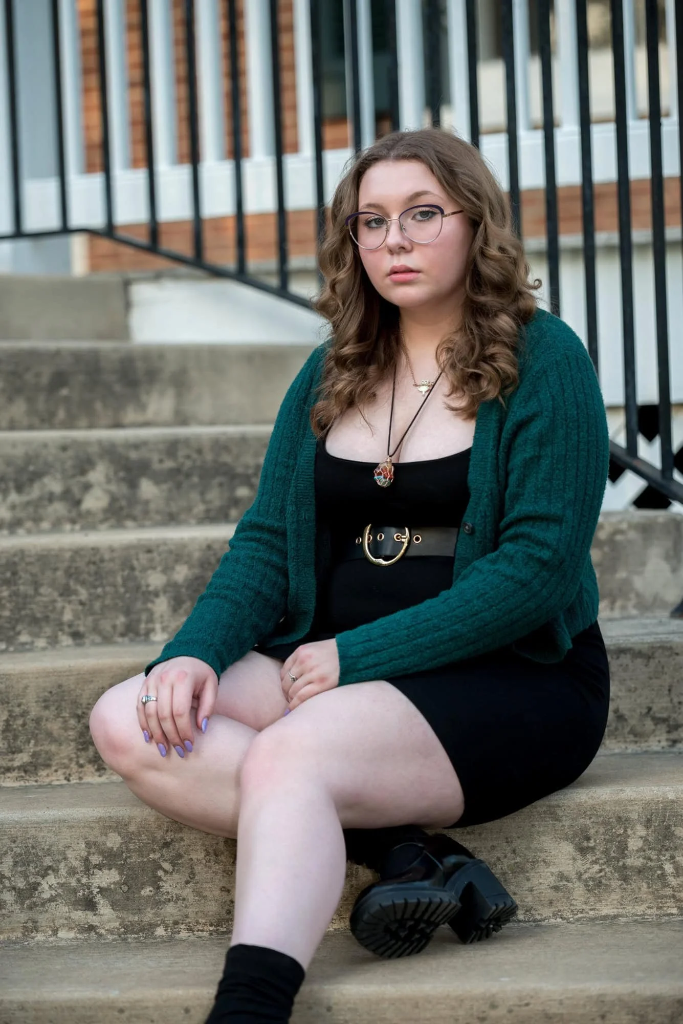 A picture captures a high school senior girl at Queeny Park wearing a black dress and green cardigan sitting on the steps of the Victorian home.