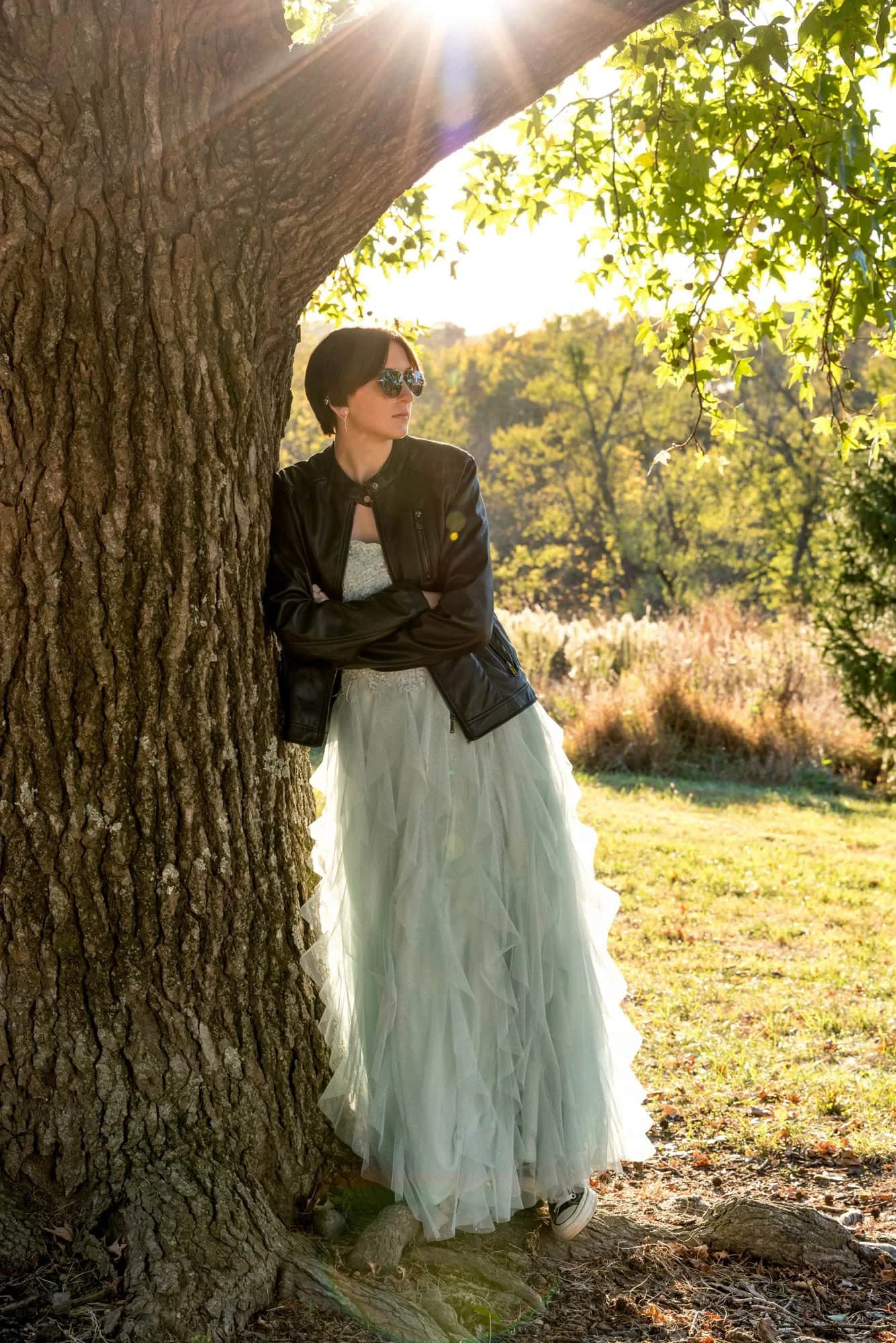 Dreamy outdoor senior photo of a girl with a serious expression crossing her arms in a black leather jacket and mint green prom dress, leaning against a tree with a warm sunset glow at Queeny Park.