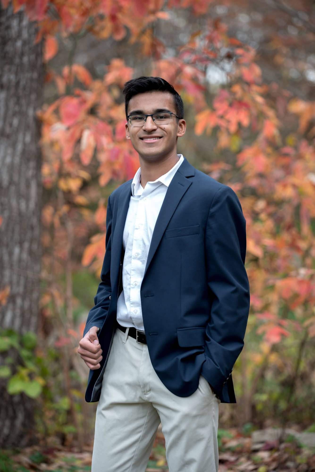 Senior portrait by a St. Louis senior portrait photographer, featuring a guy turned sideways holding his navy blazer jacket out slightly against a background of glowing orange fall leaves at Faust Park.