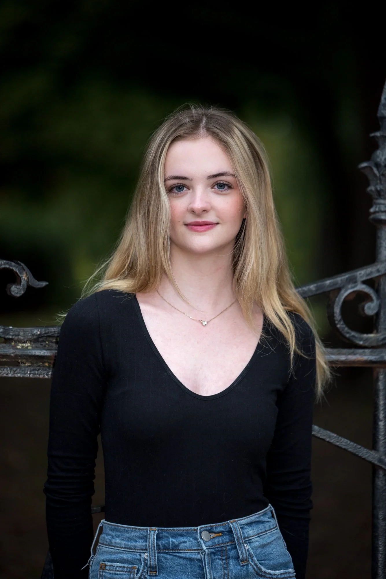 A high school senior girl in a black long-sleeve top and blue jeans leans against an iron fence at Lafayette Park for an urban style picture.