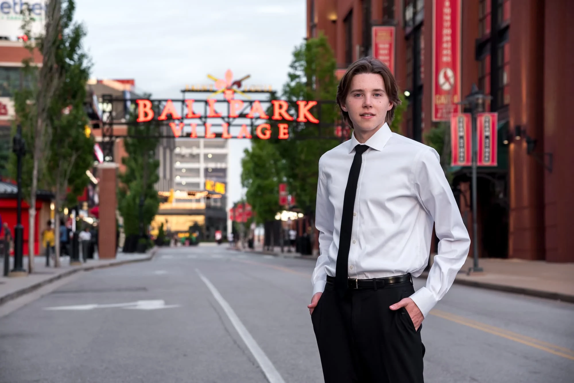 High school senior standing in the middle of a downtown street in front of the Ballpark Village sign and Busch Stadium, dressed in a sharp white long-sleeve button-up shirt, black tie, and black dress pants.