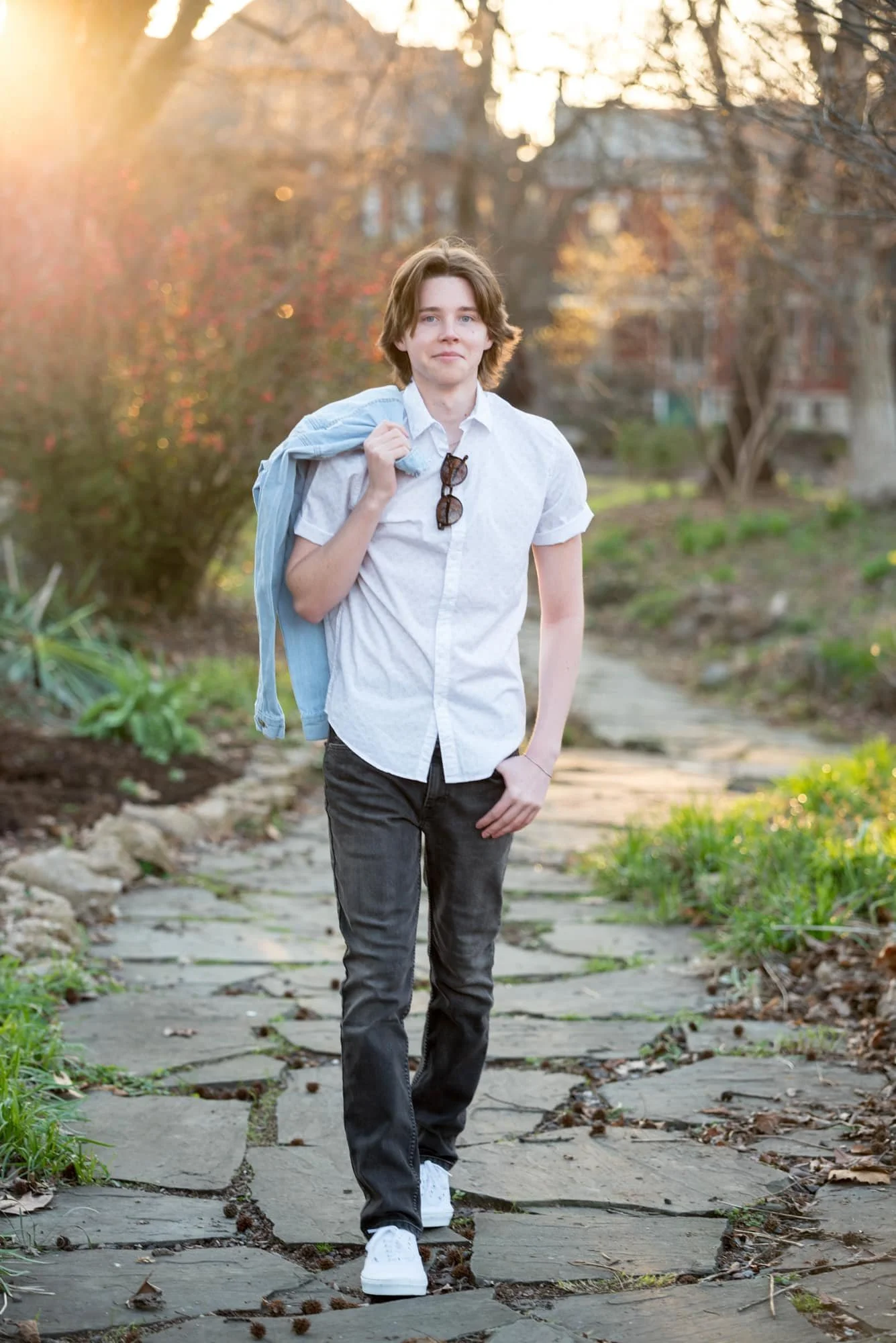 Cool, edgy senior photo of a guy walking along a stone pathway with early spring sunlight catching his sunglasses, wearing black jeans and white shoes with a casual jean jacket thrown over his shoulder at Lafayette Park.