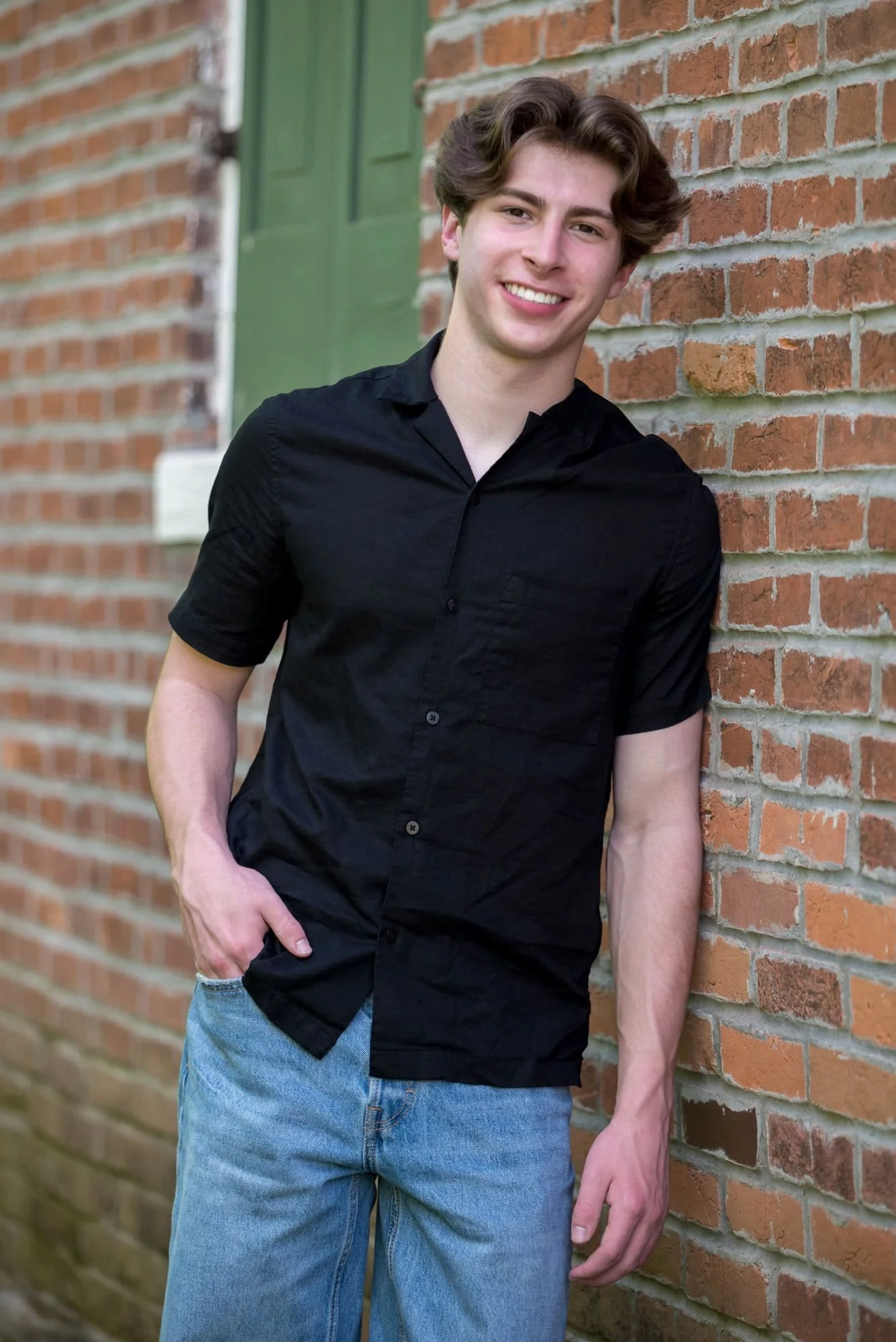 A picture captures a high school senior boy wearing a black shirt and blue jeans leaning against a brick wall in Columbia, Illinois.