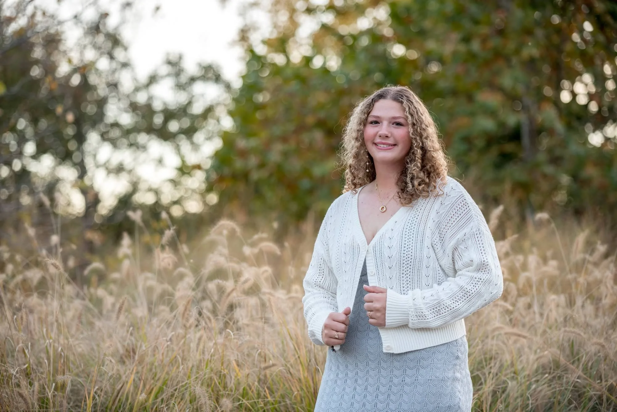 Outdoor senior picture in Forest Park of a girl posing in a white cardigan worn over a gray lace dress in front of tall ornamental grasses.
