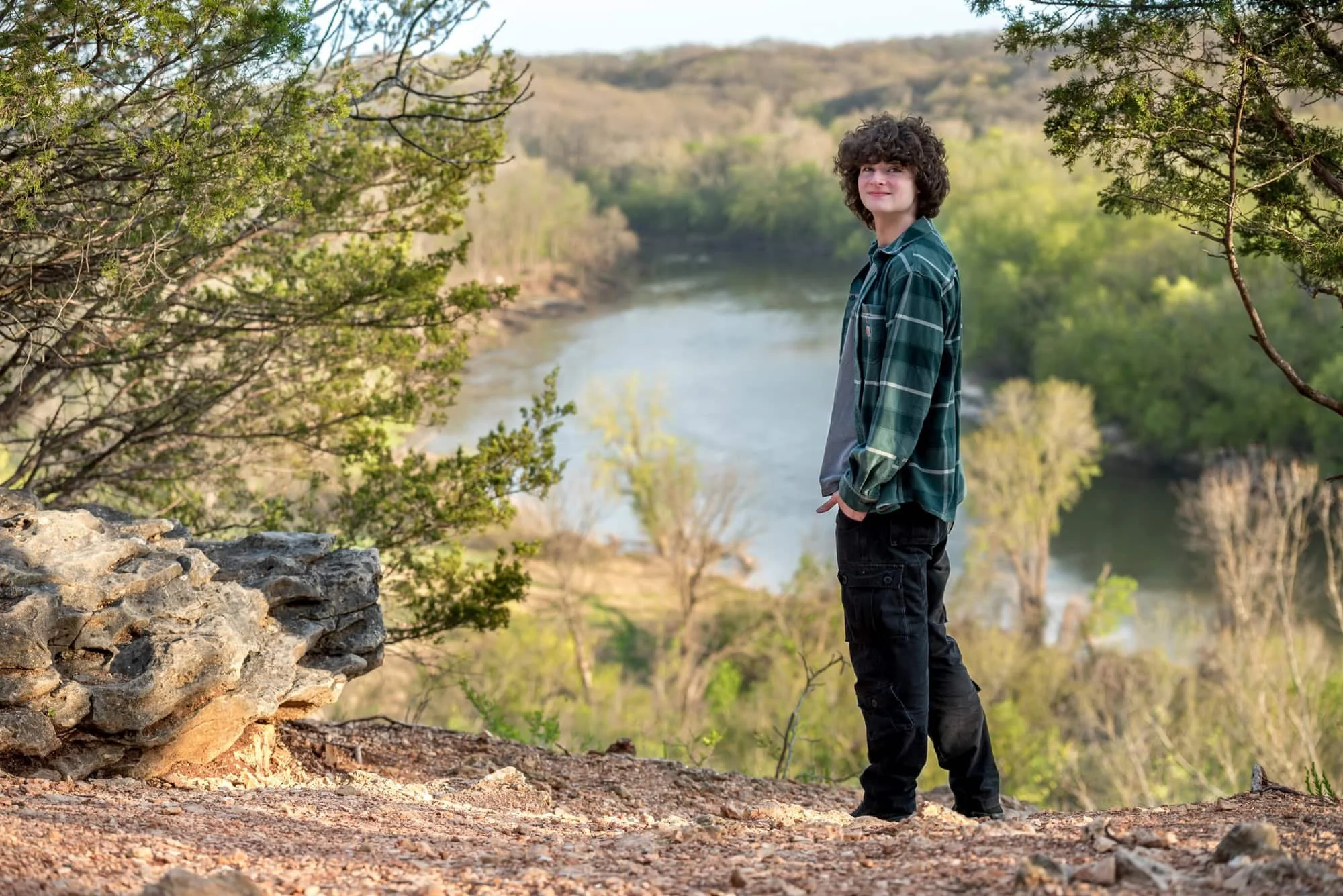 Senior picture of a guy looking back over his shoulder from a gravel overlook bluff at sunset, surrounded by sunlit spring trees and rocks with the river in the distance for a rugged West County senior photo.