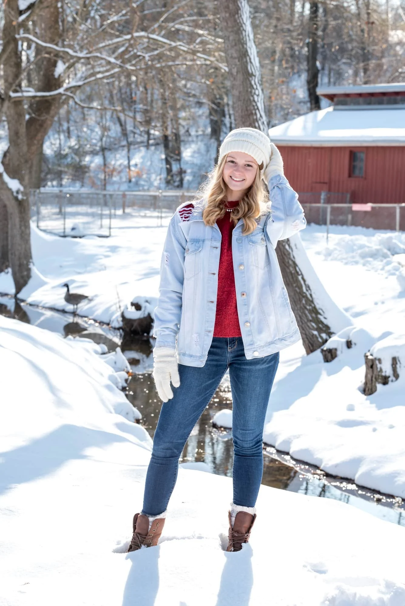 Charming country style senior portrait by an Arnold MO senior photographer, showing a girl in a light blue jean jacket, red sweater, and white beanie standing in the snow at Suson Park with a rustic red barn and geese in the background.