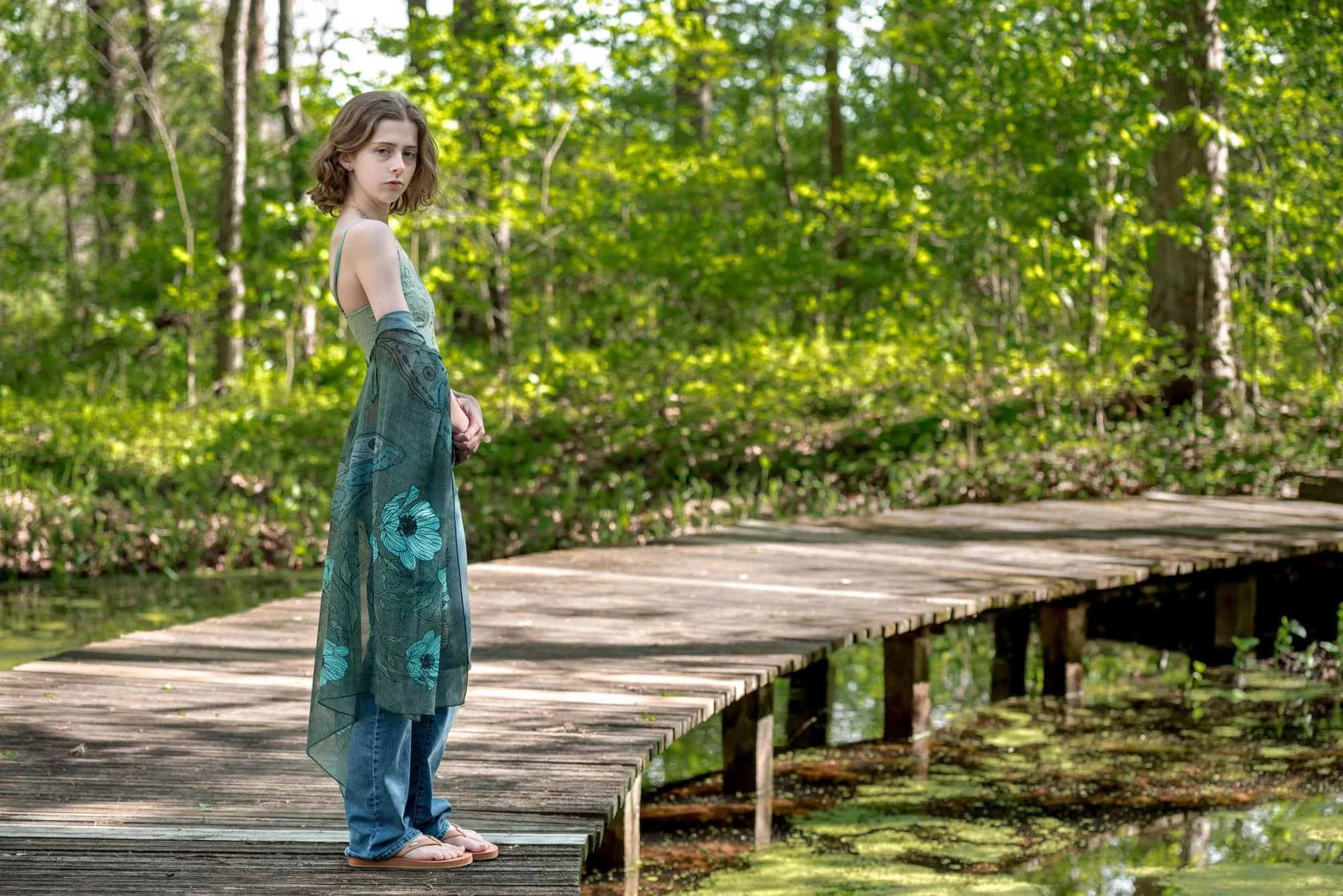 High school senior picture of a girl with a serious look standing on a wooden boardwalk over a swamp at Shaw Nature Reserve, wearing a green tank top, blue jeans, and brown flip flops with a teal floral shawl draped around her arms.