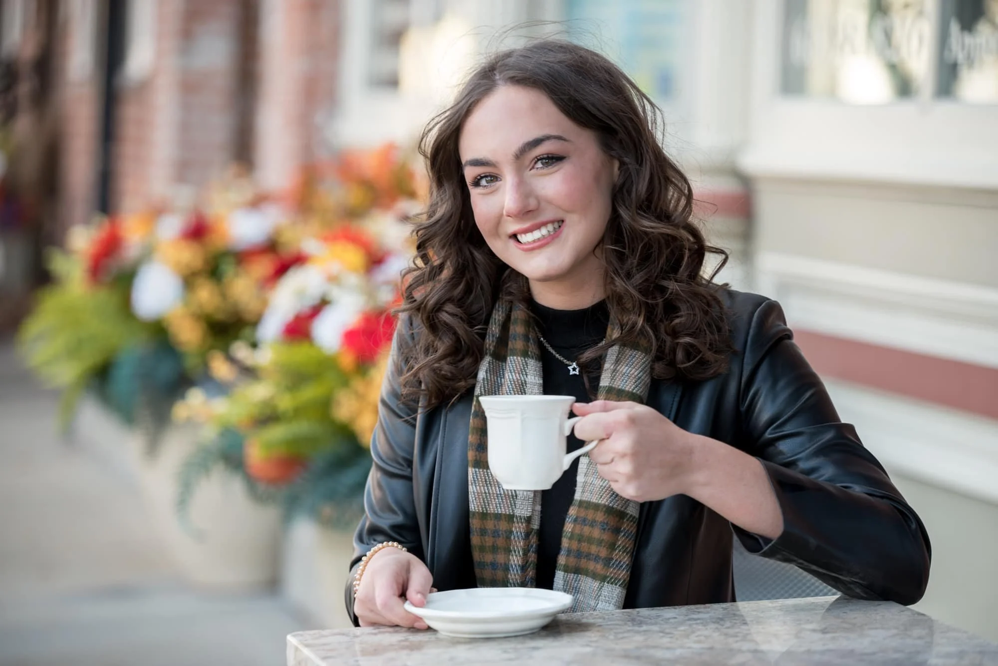 Stylish, edgy senior photo of a girl with curled hair wearing a black leather jacket and a cozy brown scarf, sitting outside historic brick buildings in Columbia IL while holding a vintage coffee mug and saucer.