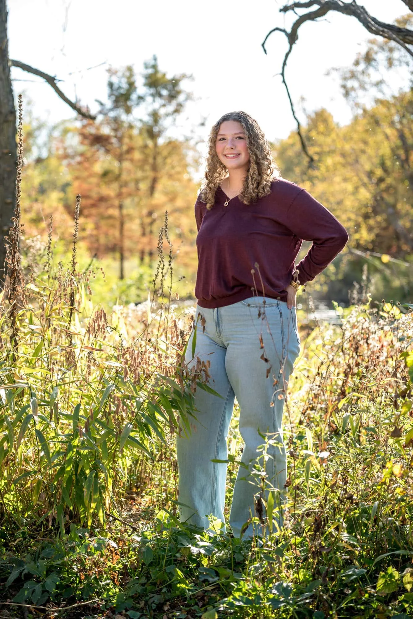 Golden hour shot taken near the Victorian Bridge showing a senior in a purple shirt standing inside a patch of sunlit wild weeds.