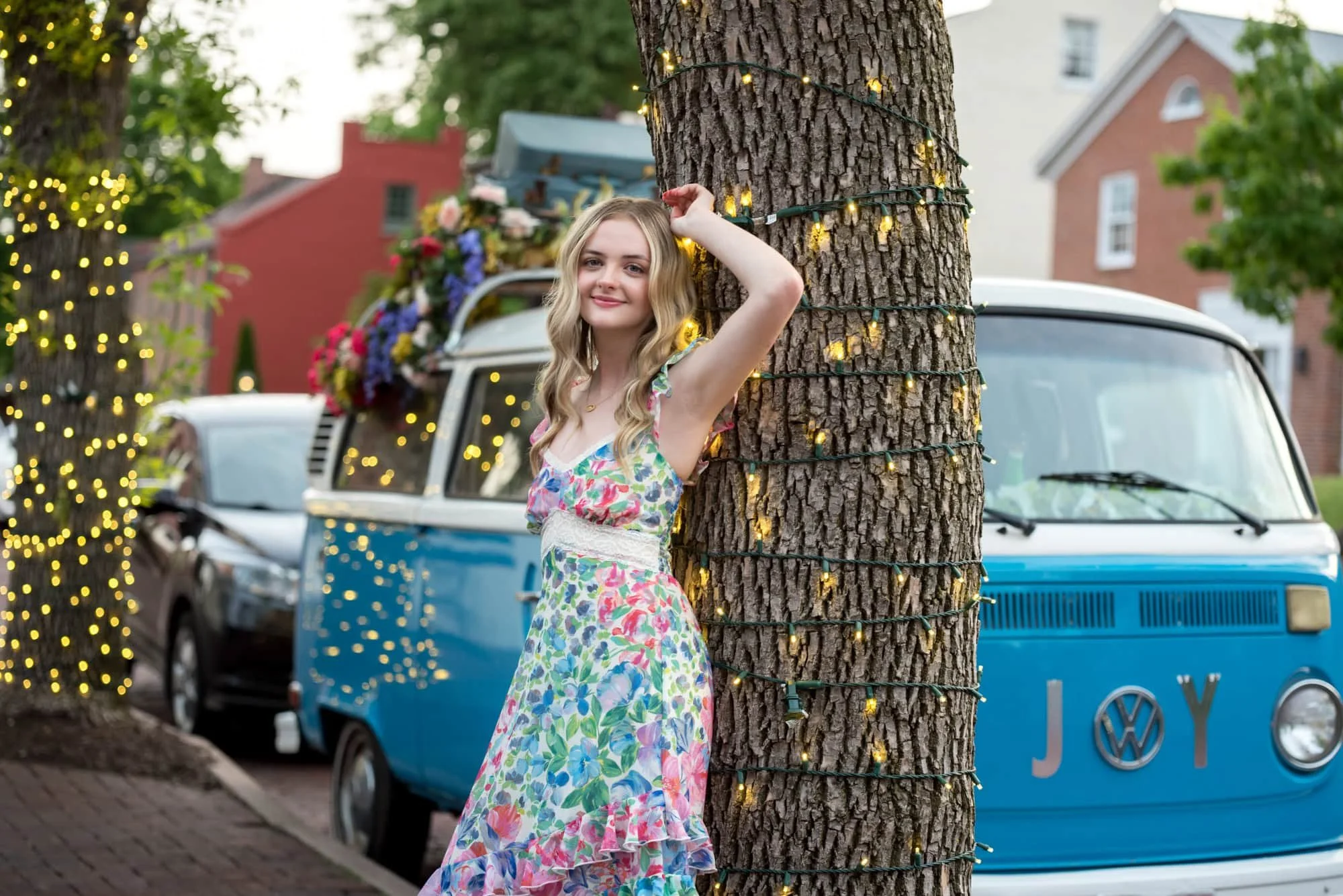 A high school senior girl leaning against a tree of lights next to a vintage VW bus on a brick sidewalk during a Main Street St. Charles senior portrait session.