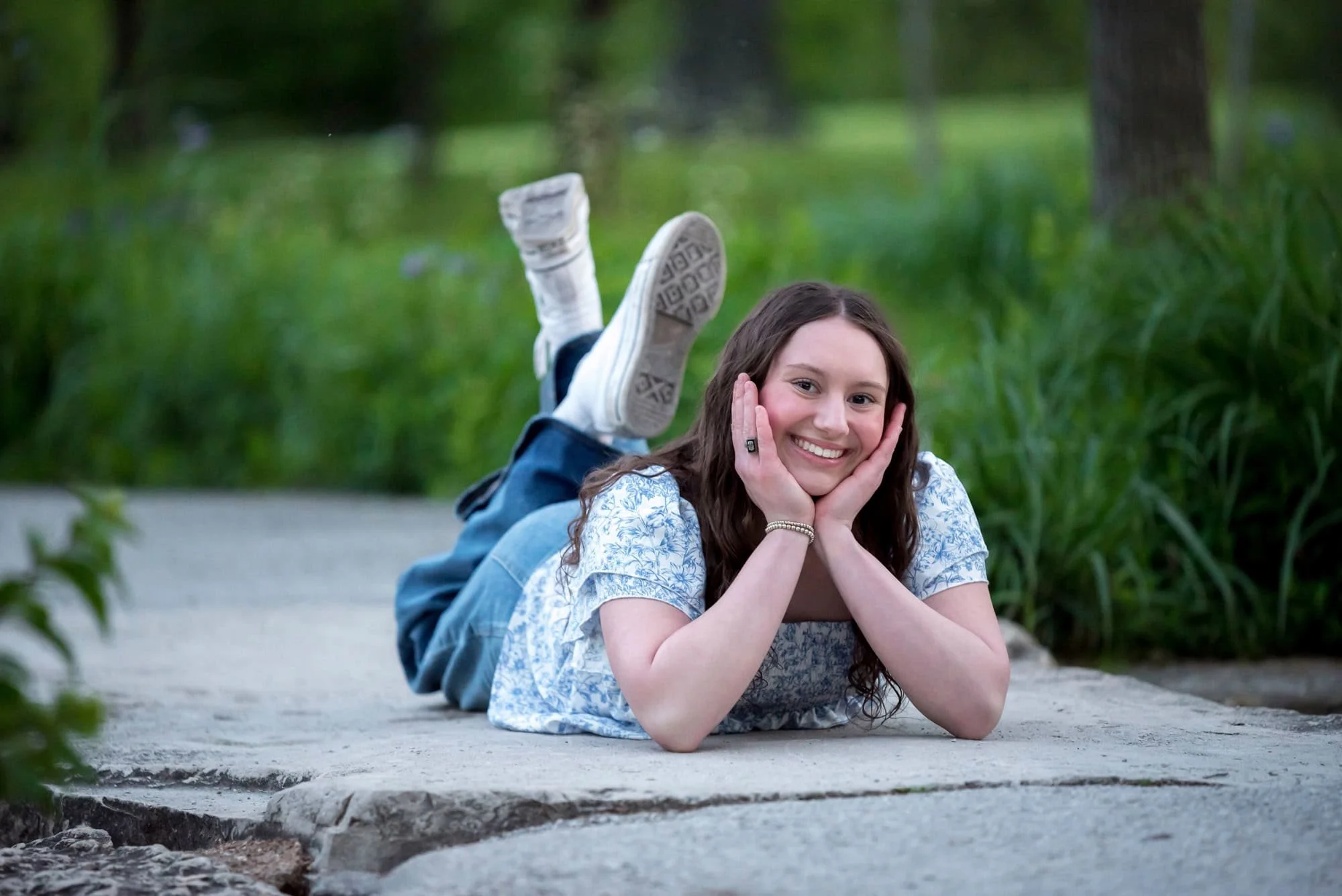 Propping her chin in her hands, a smiling senior girl lies on a stone path next to the creek with her feet kicked up behind her taken by a St. Louis senior photographer.