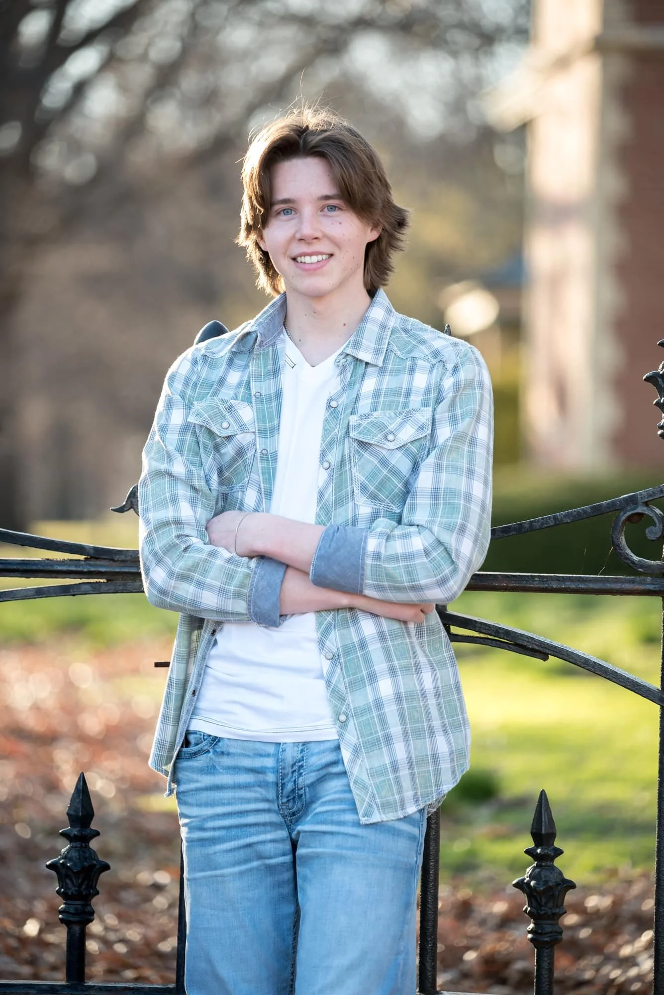 Smiling senior picture taken by a local St. Louis, Missouri photographer, showing a boy with his arms crossed leaning against a wrought iron fence with a historic brick park building and greenery in the background.