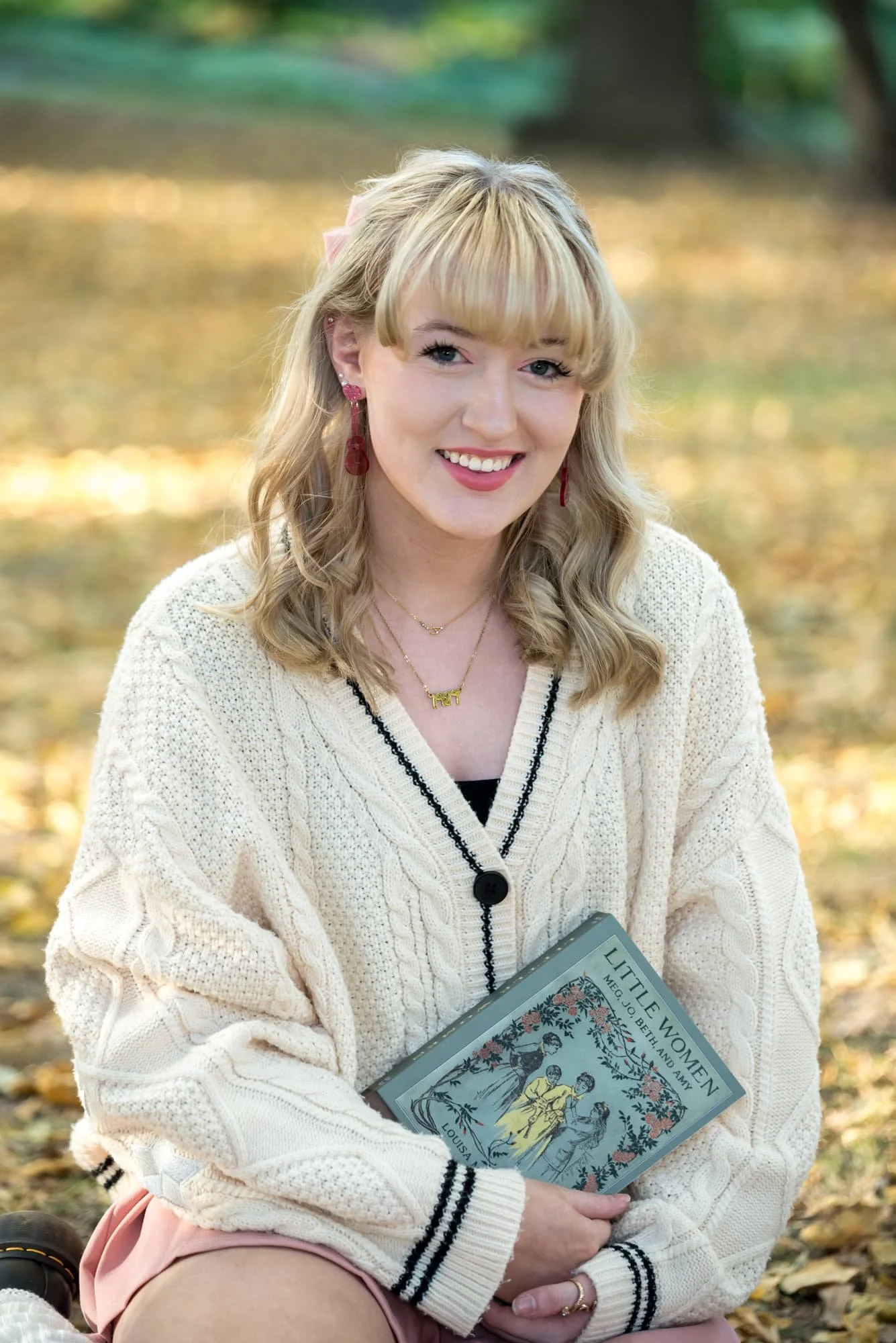 Sitting outdoors in the leaves during a fall session in St. Louis, Missouri, a senior girl holds a book for a picture in a white sweater and pink skirt.