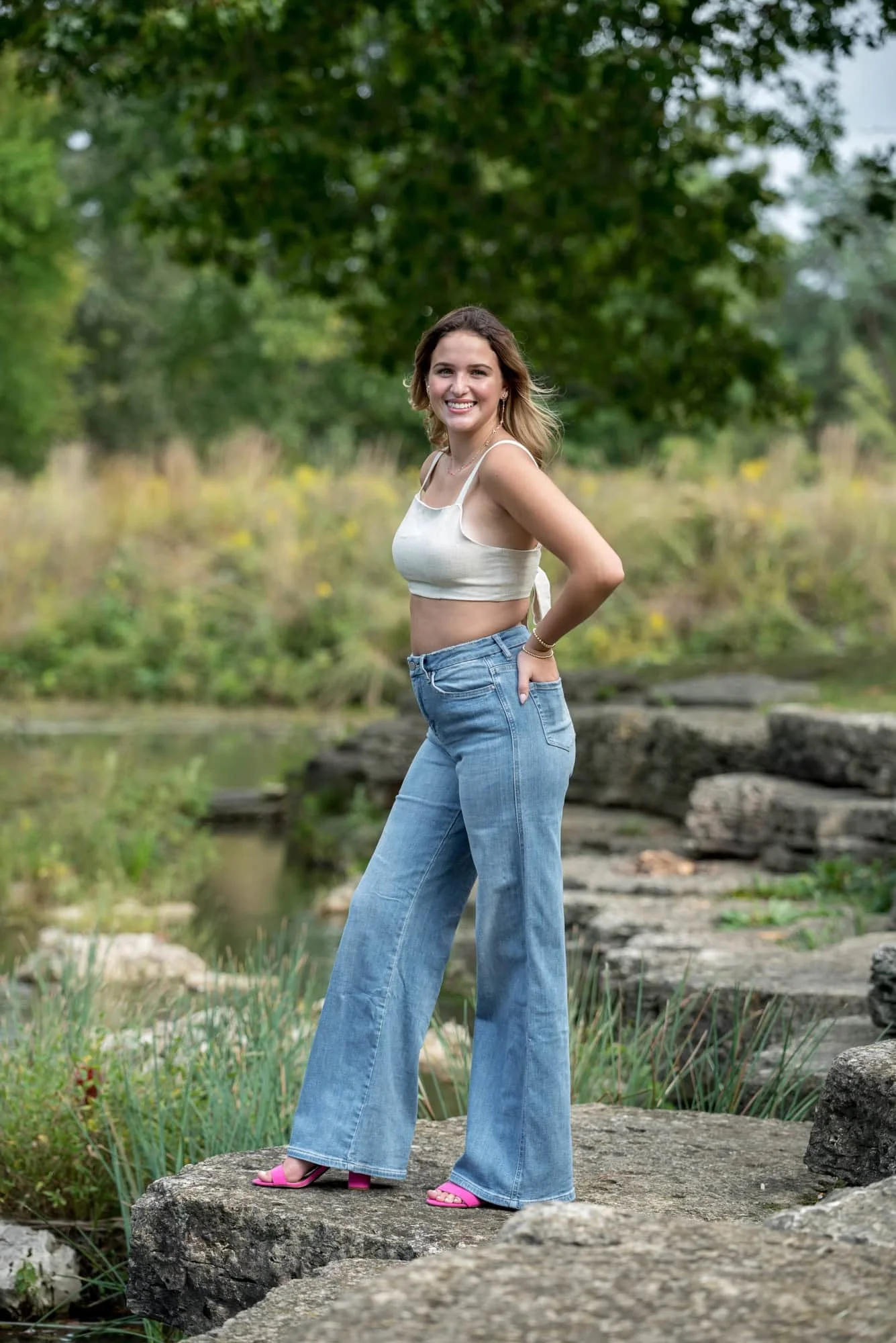 Country style creek-side photo capturing a senior wearing a tan crop top, blue jeans, and pink heels on a rock near The Muny taken by a St. Louis photographer.