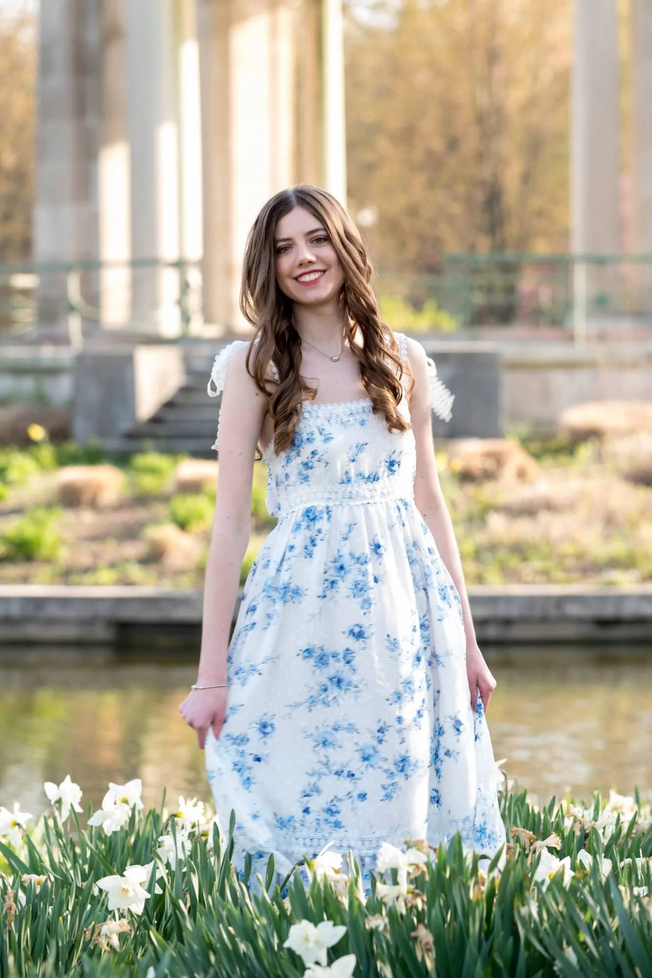 Senior girl in a white dress with blue flowers, twirling joyfully in a field of white daffodils in front of the historic Muny pagoda and pond in Forest Park.