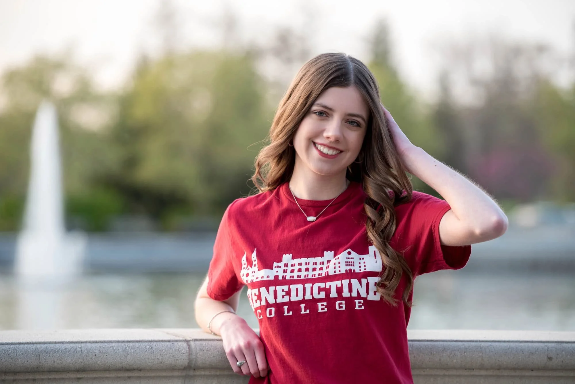 Senior girl wearing a red college shirt, leaning against a bridge with her hand in her hair and the scenic Art Hill lake basin stretching out behind her.