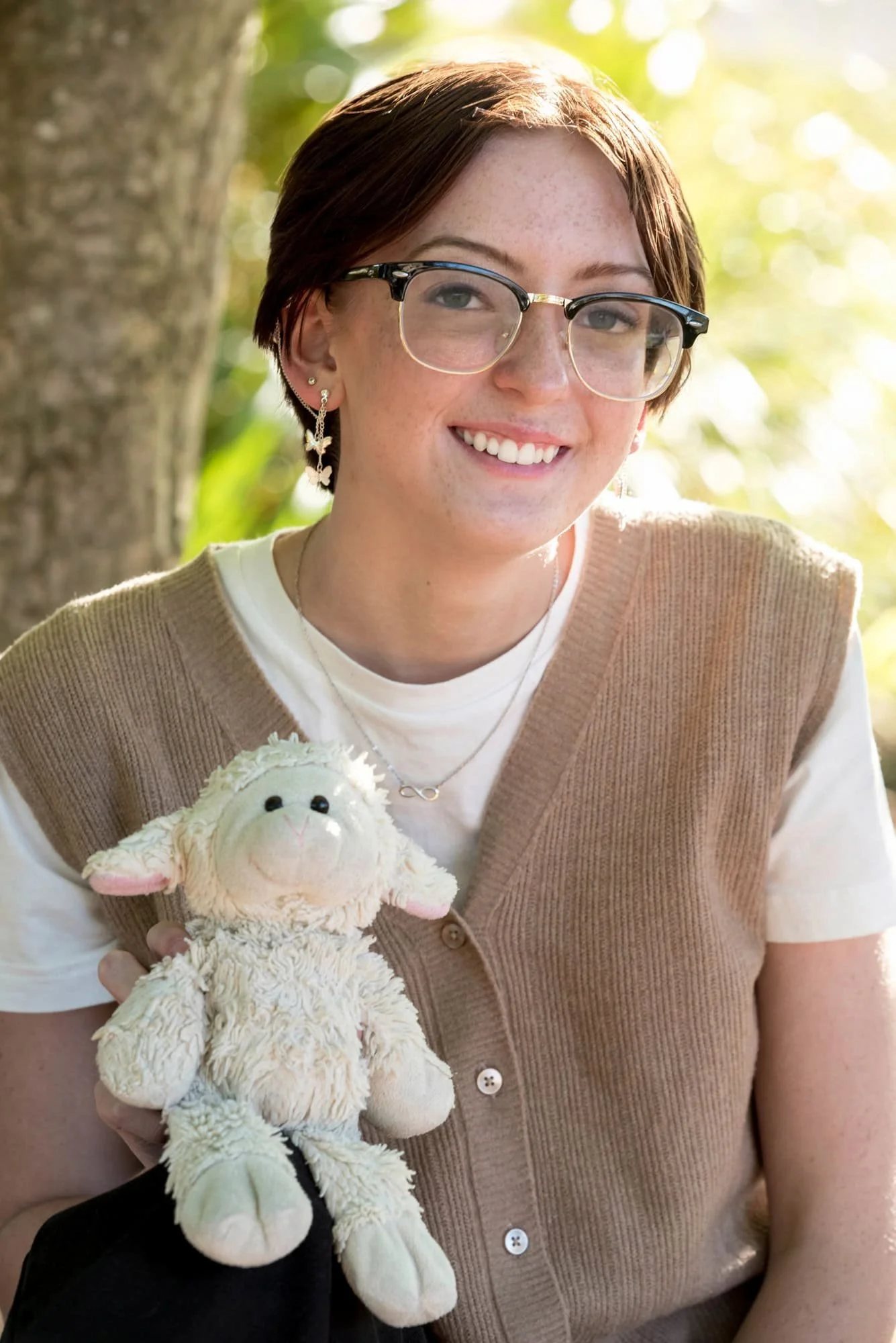 Heartwarming close-up senior picture of a girl wearing a tan vest, white t-shirt, and black jeans, sitting in front of a sunlit bush while holding her childhood stuffed lamb.