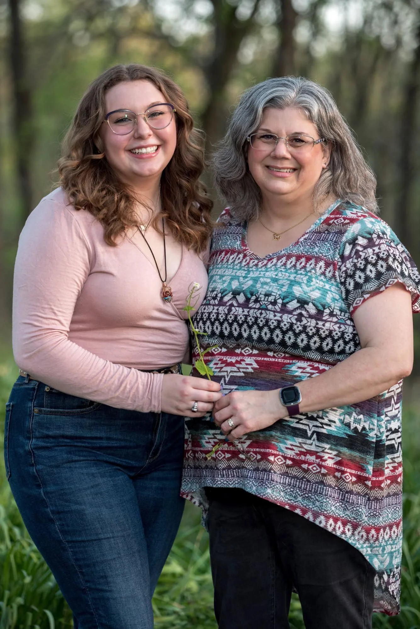 A picture captures a high school senior girl in a pink long-sleeve shirt and blue jeans standing with her mom for a photo by a Town and County photographer.
