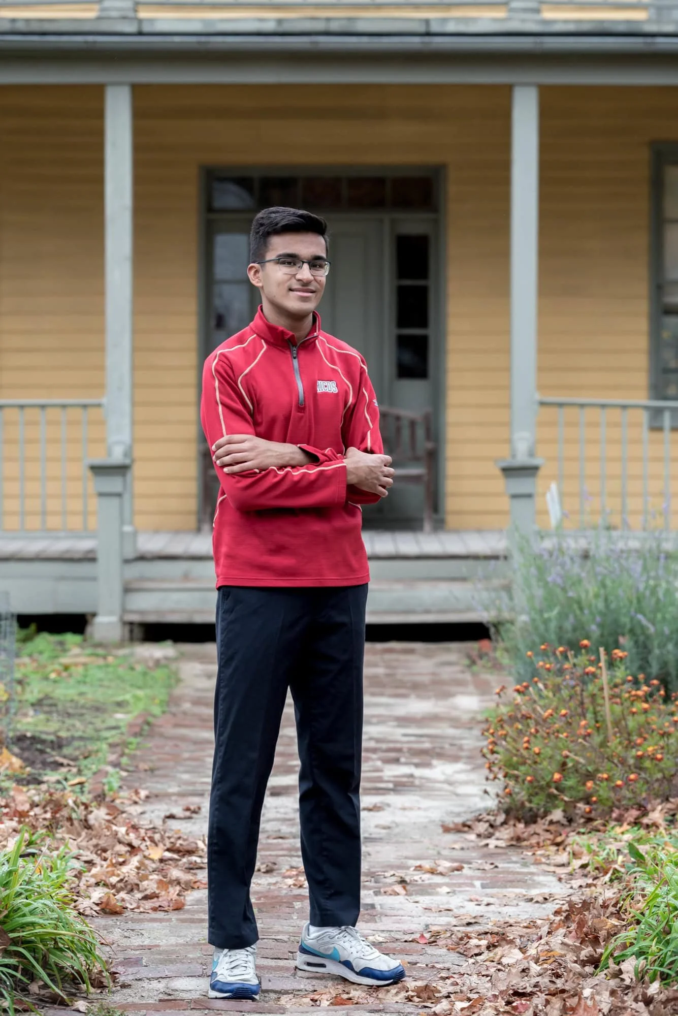 Casual senior picture of a guy with arms crossed, standing on a brick pathway in front of a historic yellow house at Faust Park in Chesterfield.