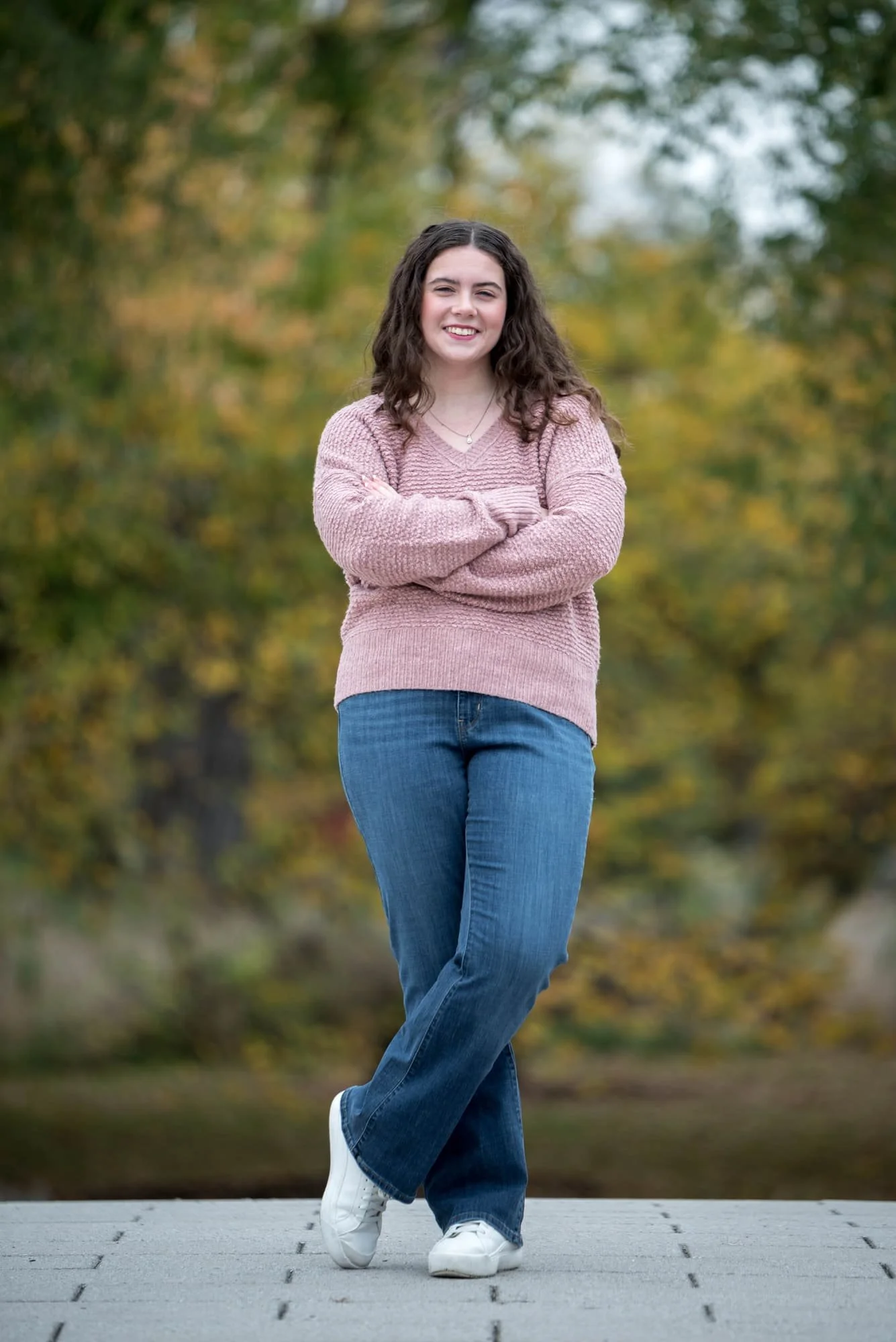 On a footbridge near Art Hill in Forest Park, a picture of a high school senior girl wearing a pink sweater and blue jeans stands in front of fall trees.