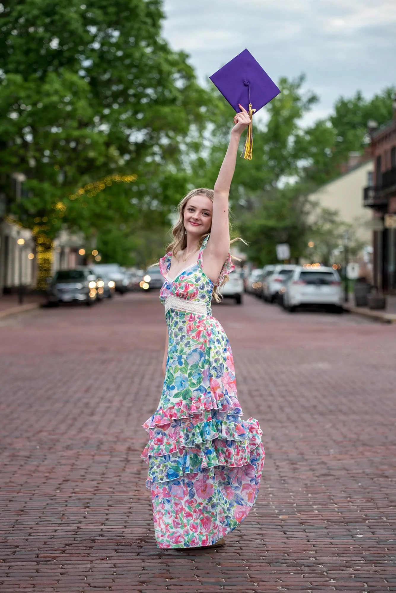 A spring photo captures a high school senior girl in a floral long dress standing in the middle of a brick road in St. Charles holding up her graduation cap.