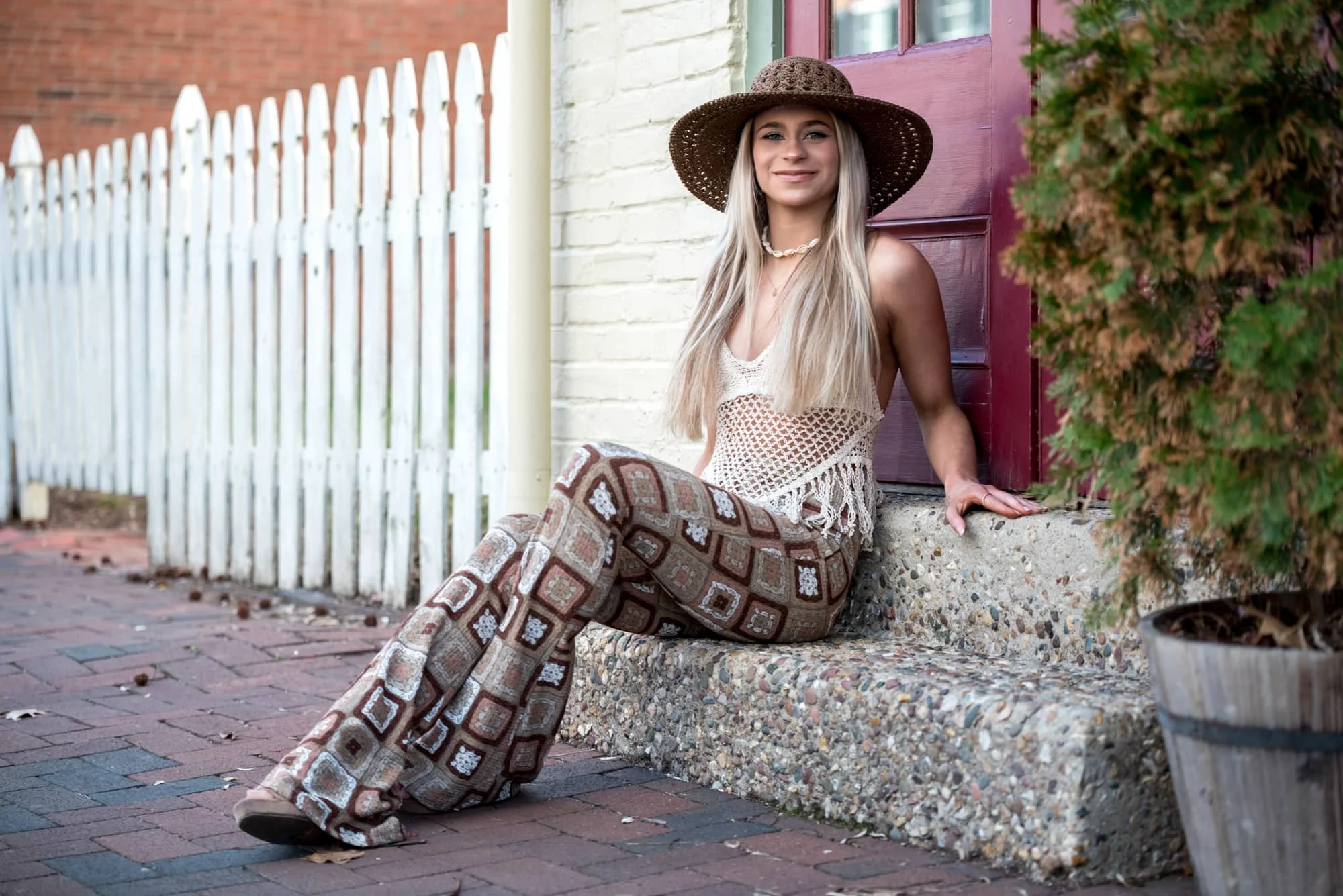 Boho style senior portrait of a girl wearing a tan floppy hat, tan crocheted crop top, and brown patterned square flare pants, sitting on historic stone steps by a red door and white fence on a brick pathway in St. Charles MO.