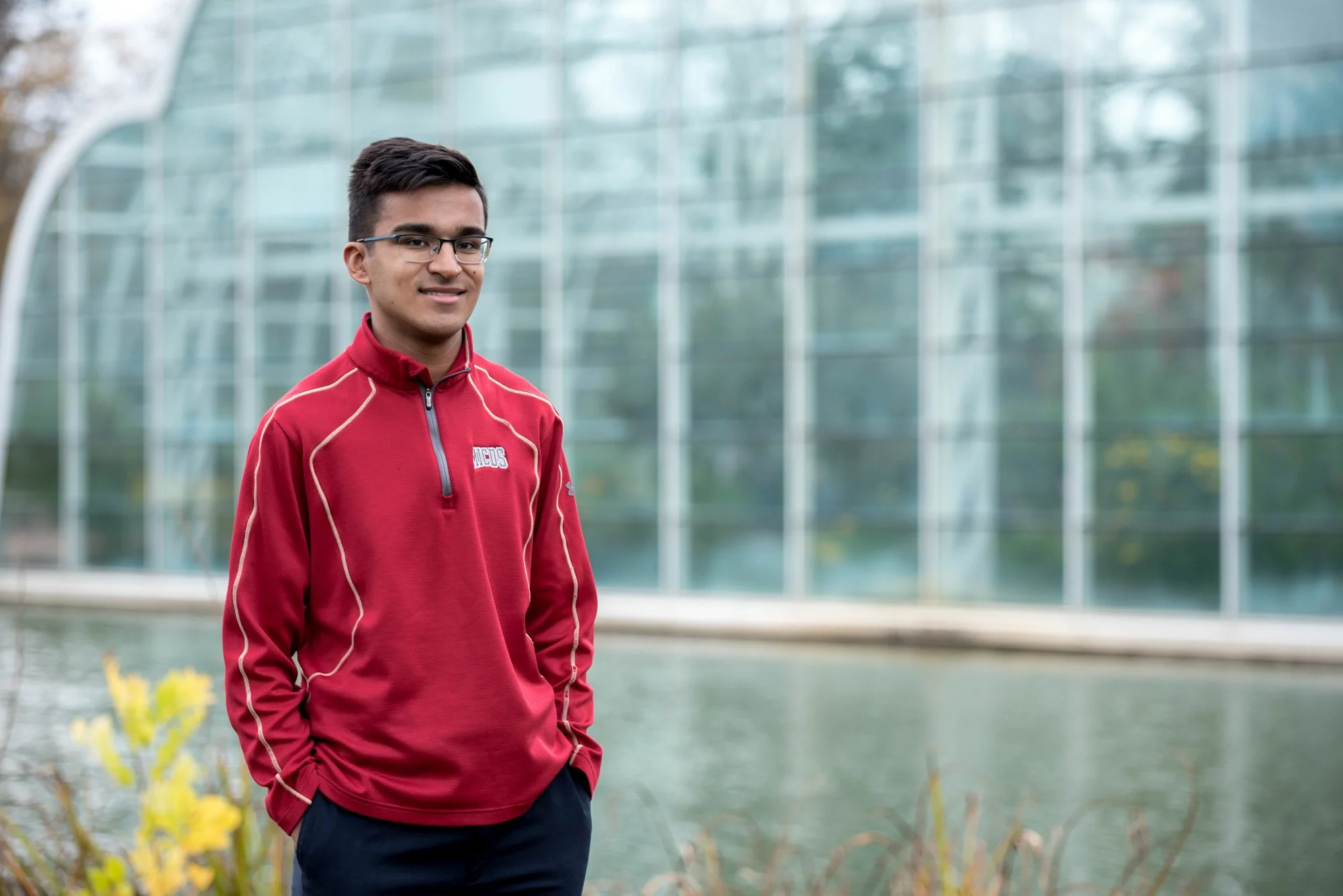 Senior picture by a Chesterfield MO senior photographer, featuring an Indian boy in a red zip-up school shirt and black pants standing with his hands in his pockets in front of the Sophia M. Sachs Butterfly House and pond.