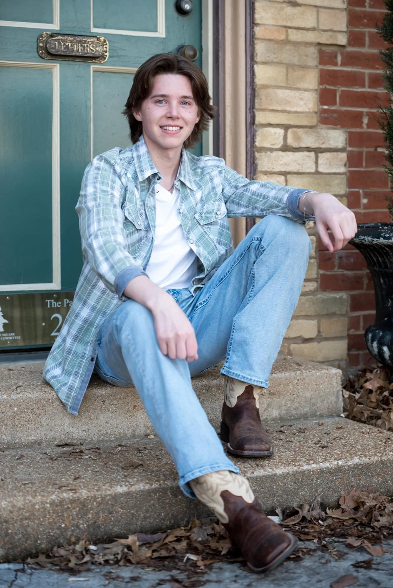 Casual high school senior portrait of a guy wearing a blue and green plaid shirt over a white tee, blue jeans, and cowboy boots, sitting relaxed on the historic stone steps in front of a building with a green door at Lafayette Park.