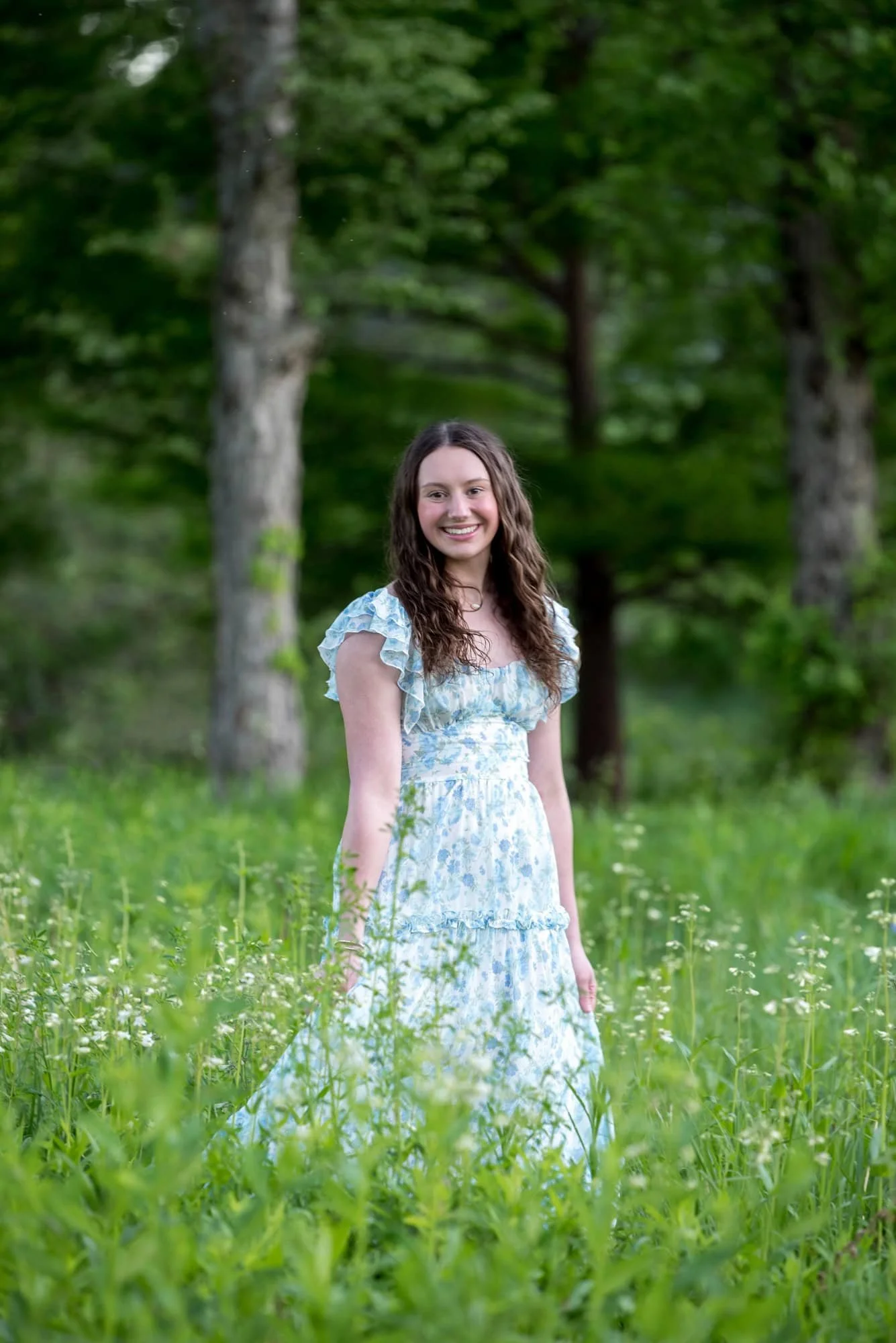 High school senior girl in a light blue floral dress smiling while twirling in a whimsical, wildflower-filled meadow in Forest Park.