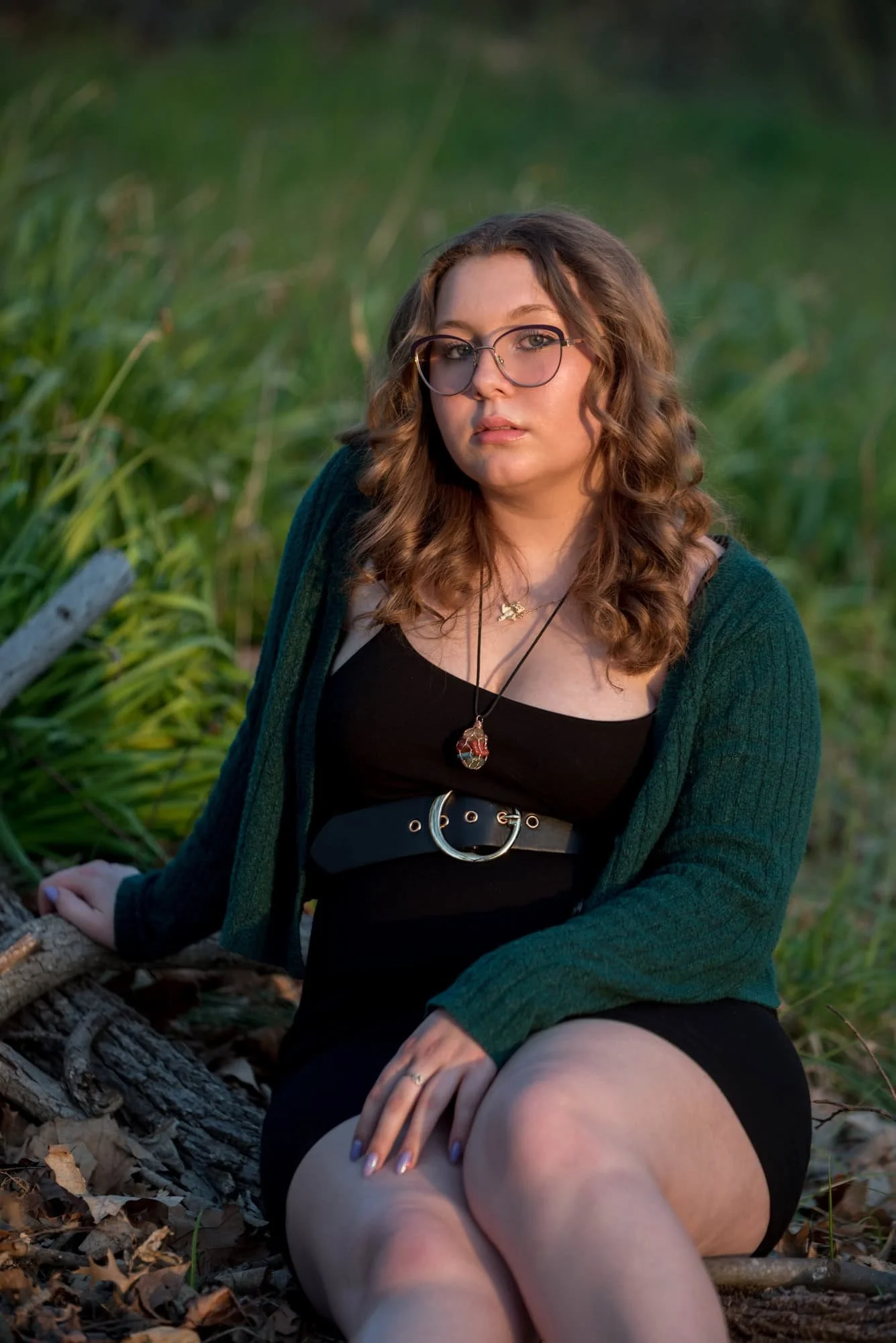 A picture captures a high school senior girl wearing a black dress and green cardigan in a wooded area of Queeny Park during sunset.