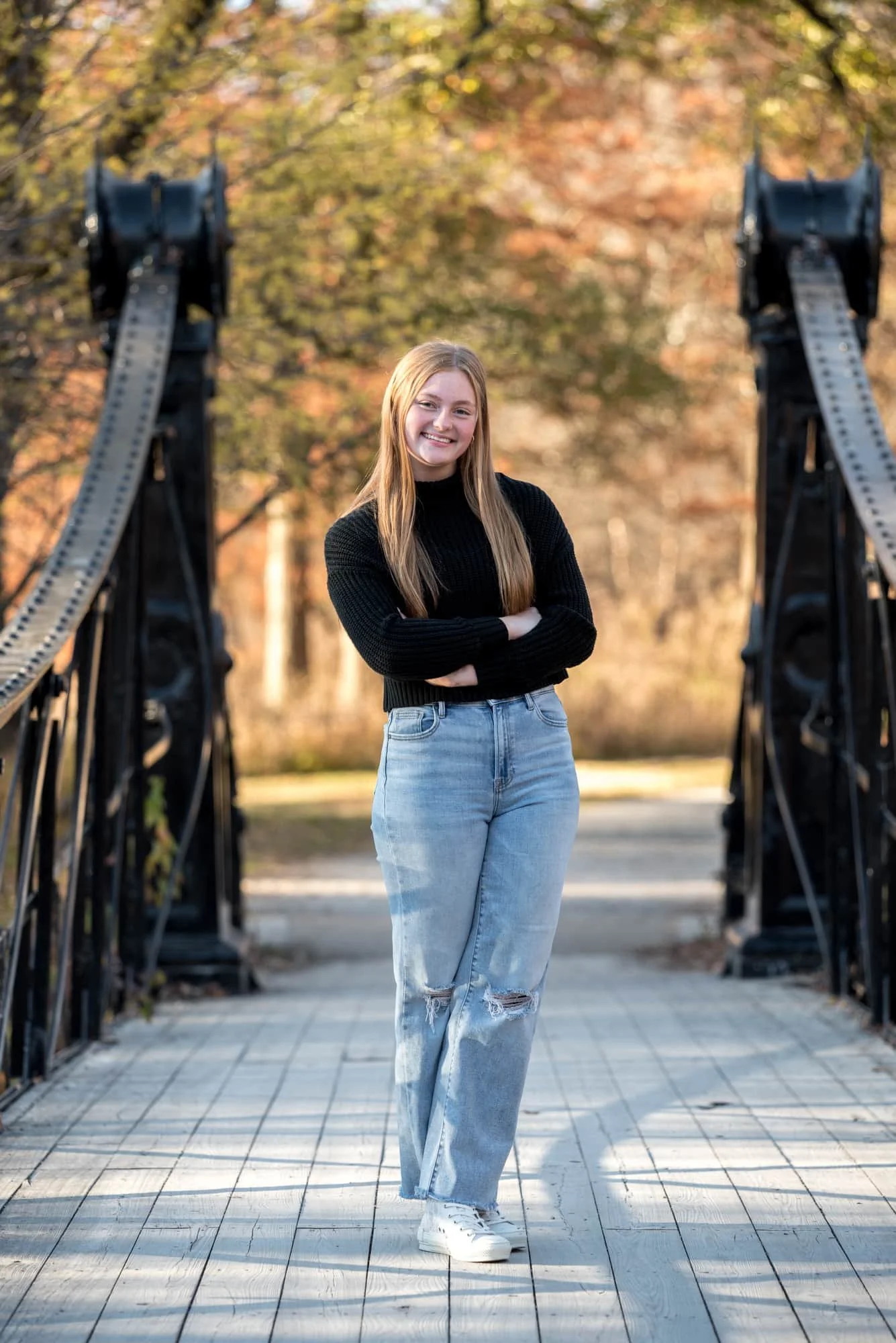 A fall picture shows a high school senior girl wearing a black sweater and blue jeans standing on the Victorian Bridge.