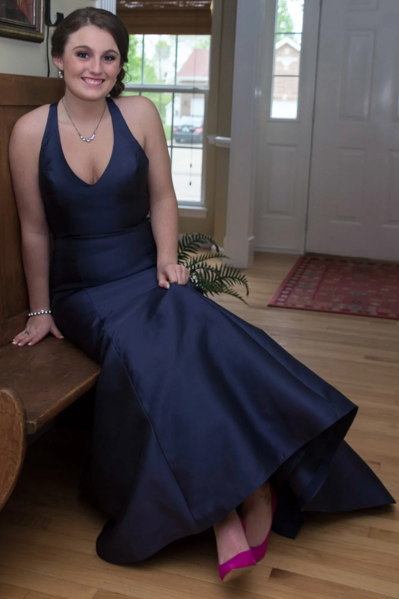 Picture of a girl smiling while sitting on a rustic wooden bench inside a home in Monroe County, Illinois, showing off her navy blue prom gown and vibrant hot pink high heels.