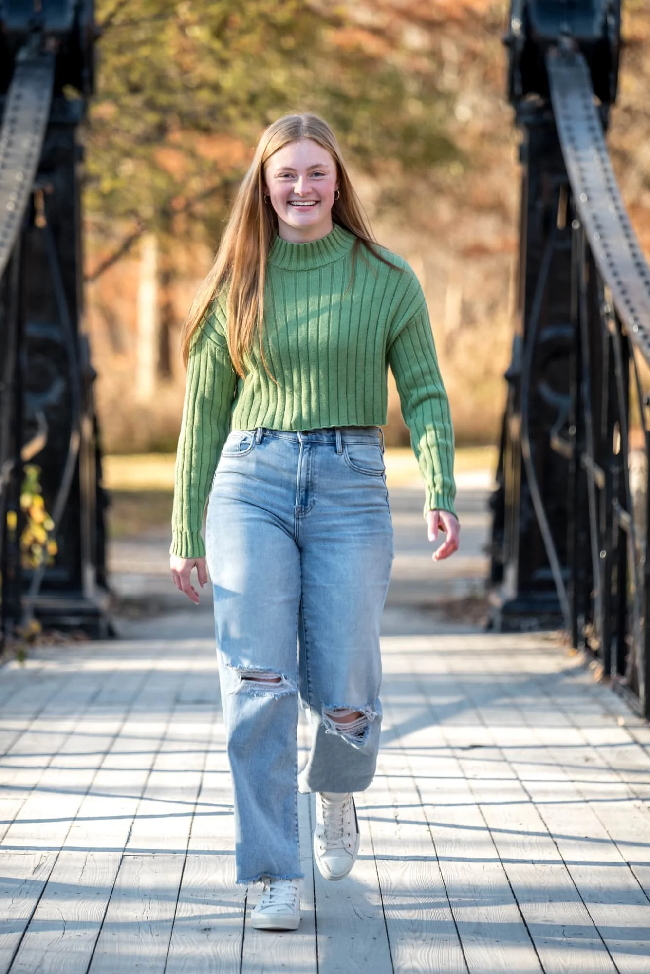 A fall photo captures a high school senior girl wearing a green sweater and blue jeans walking across  the Victorian Bridge in Forest Park.