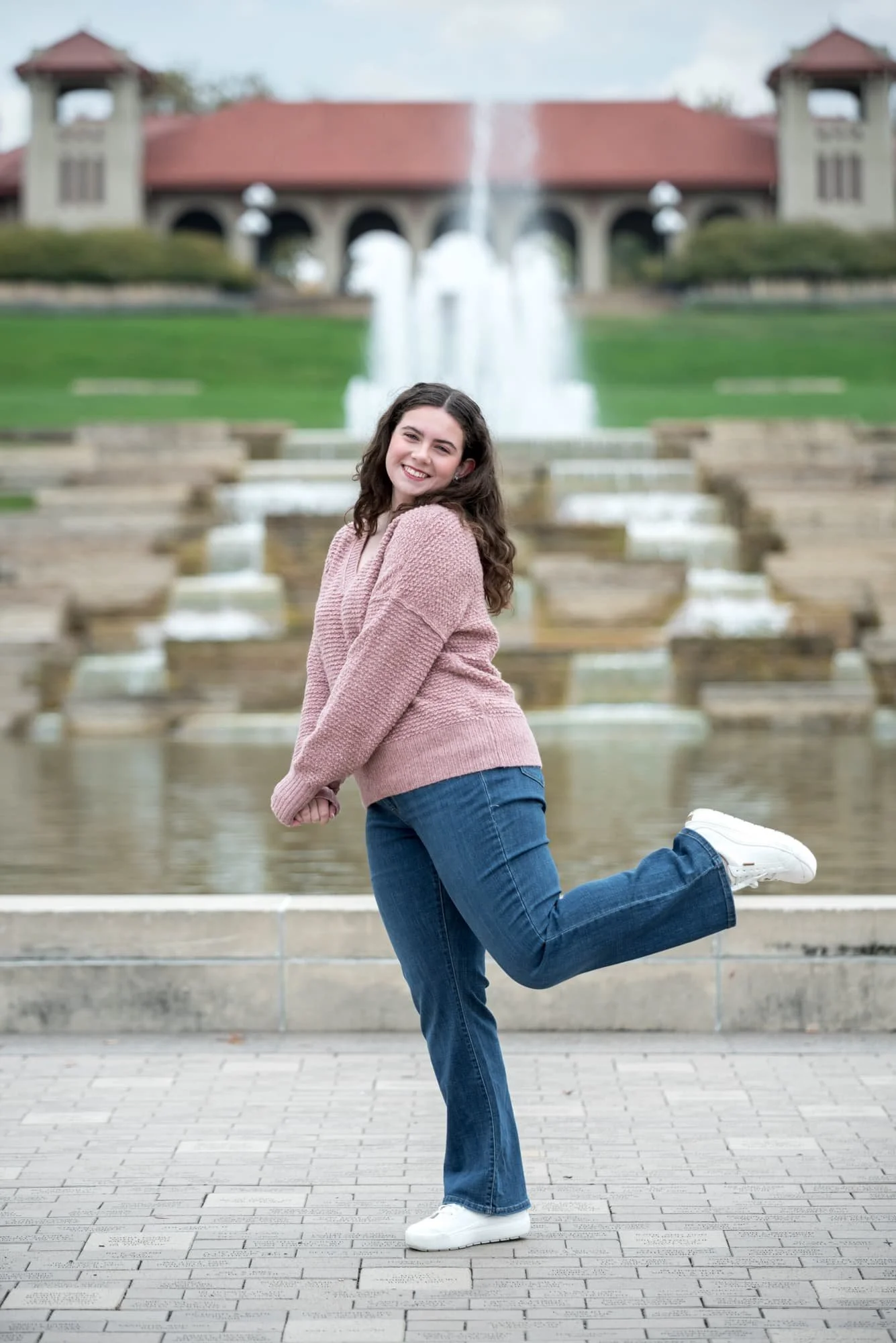 Charming senior photo of a girl wearing a light pink sweater, blue jeans, and white shoes, posing with her hands clasped and one leg kicked up in front of the grand water fountain pool at the World's Fair Pavilion base in Forest Park.