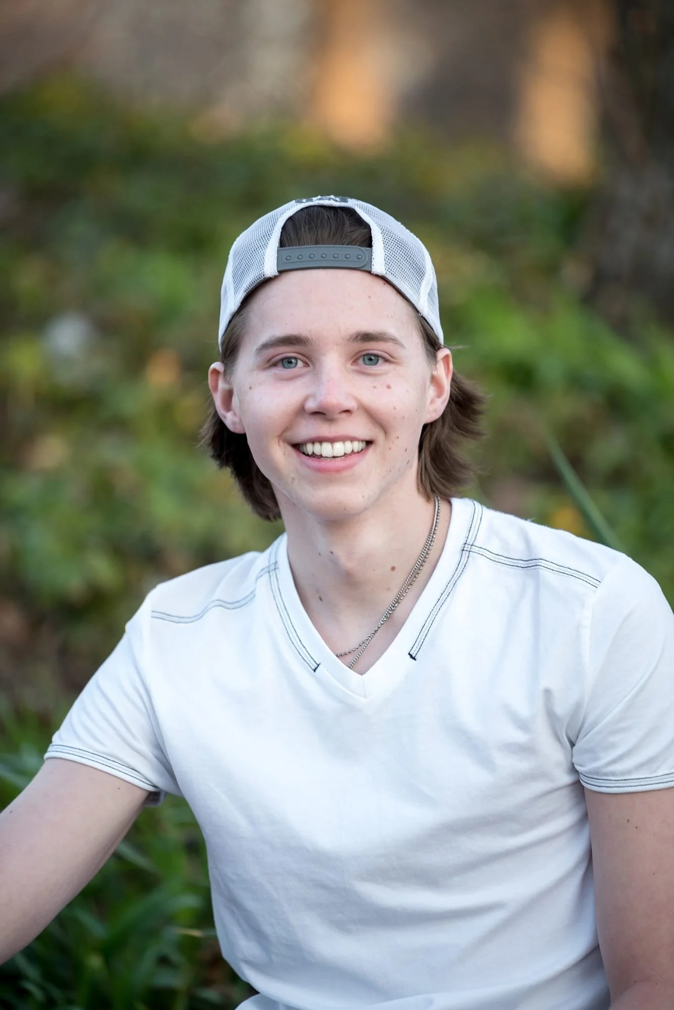 Close-up senior pphoto taken at in St. Louis, MO, showcasing a young man wearing a backwards baseball cap and a white t-shirt against a background of green park shrubbery.