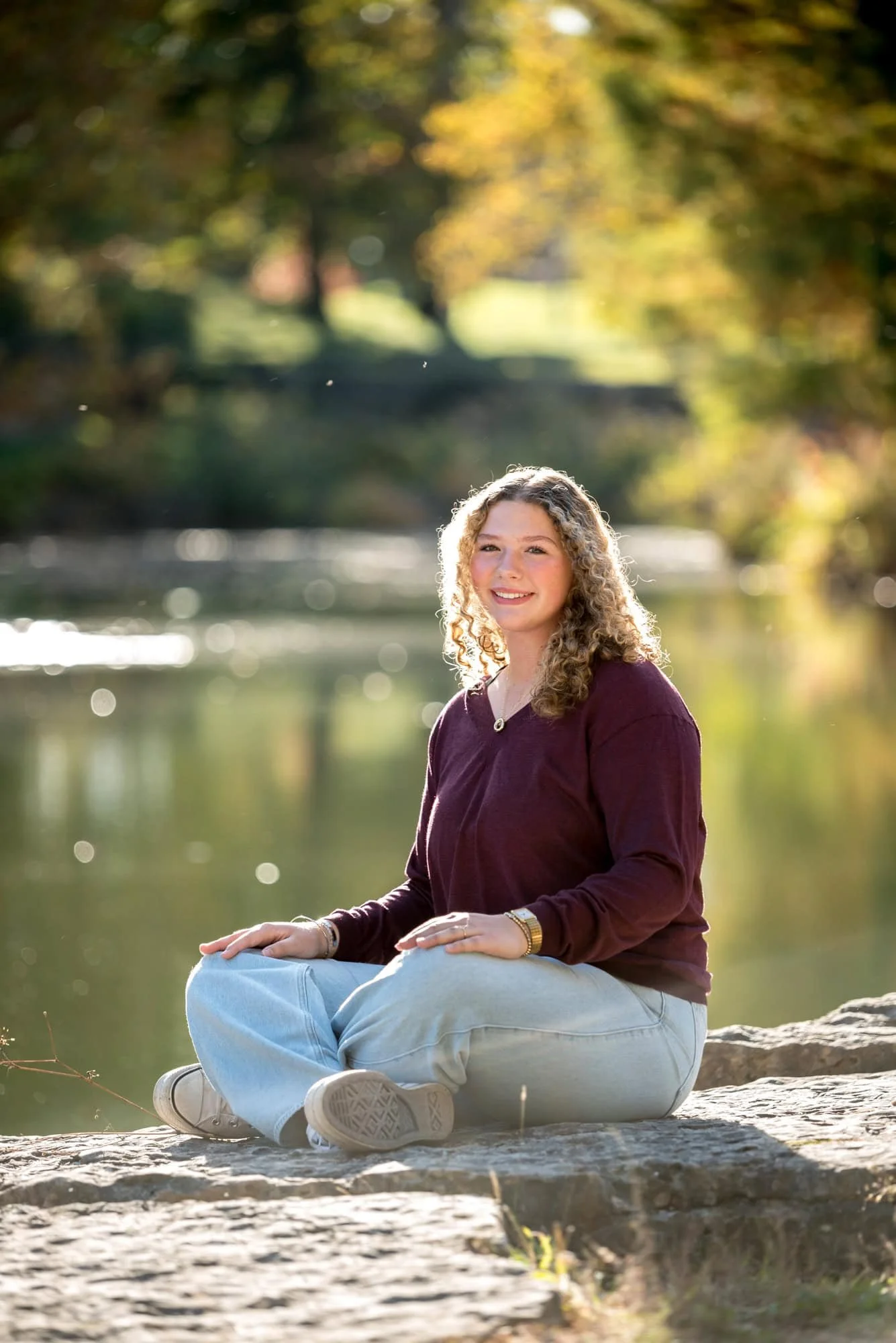 Natural country style photo in St. Louis of a girl wearing a purple shirt and blue jeans sitting cross-legged beside a woodland creek.