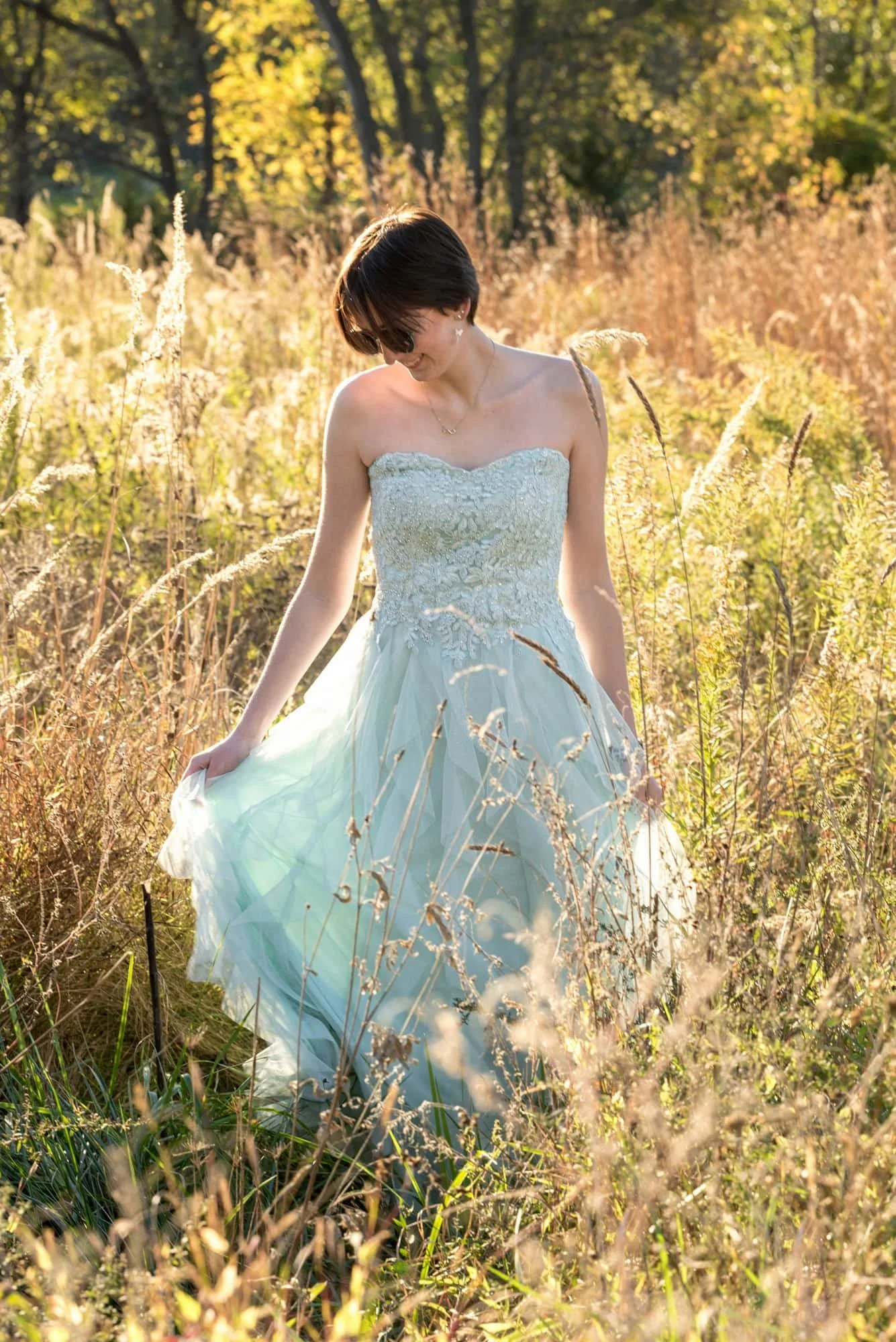 Whimsical senior picture taken by a St. Louis senior photographer, capturing a girl wearing sunglasses and twirling around in a mint green prom dress in a sunlit field of tall grass at Queeny Park in St. Louis County.