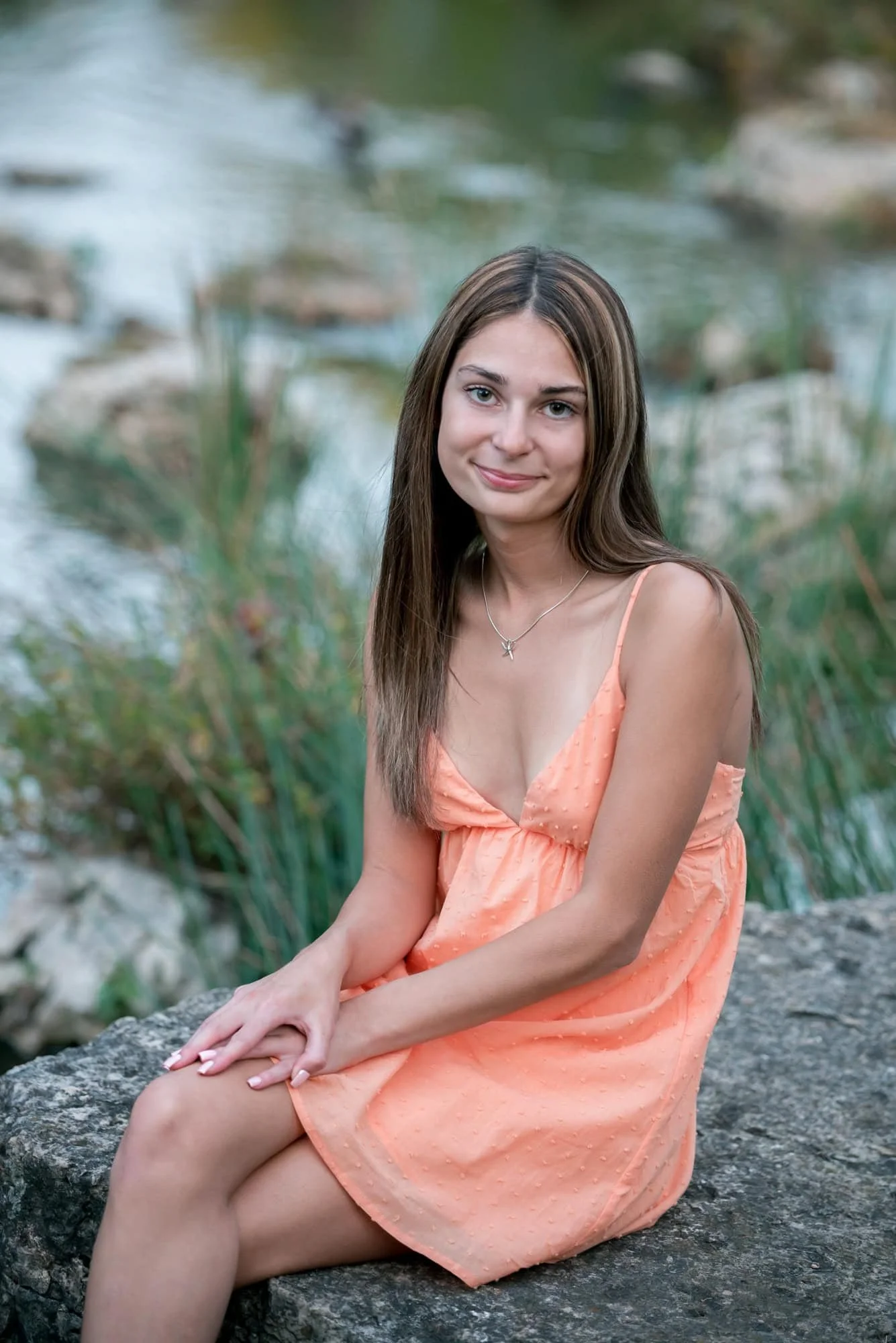 Natural country style photo near the Muny showing a senior in an orange dress sitting on a rustic creek-side rock in the region.