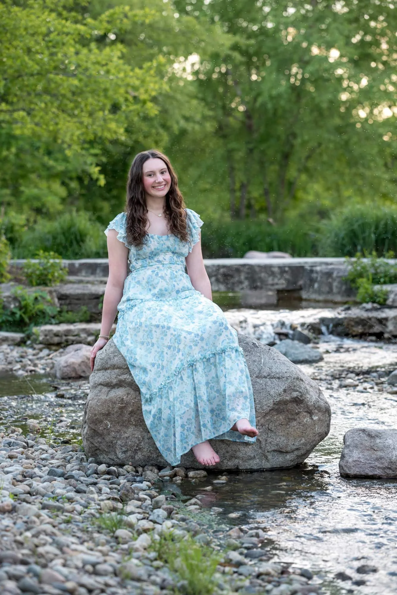 Barefoot and smiling, a senior girl sits on a large boulder in the middle of a flowing, stone-filled country creek during her session near Art Hill.