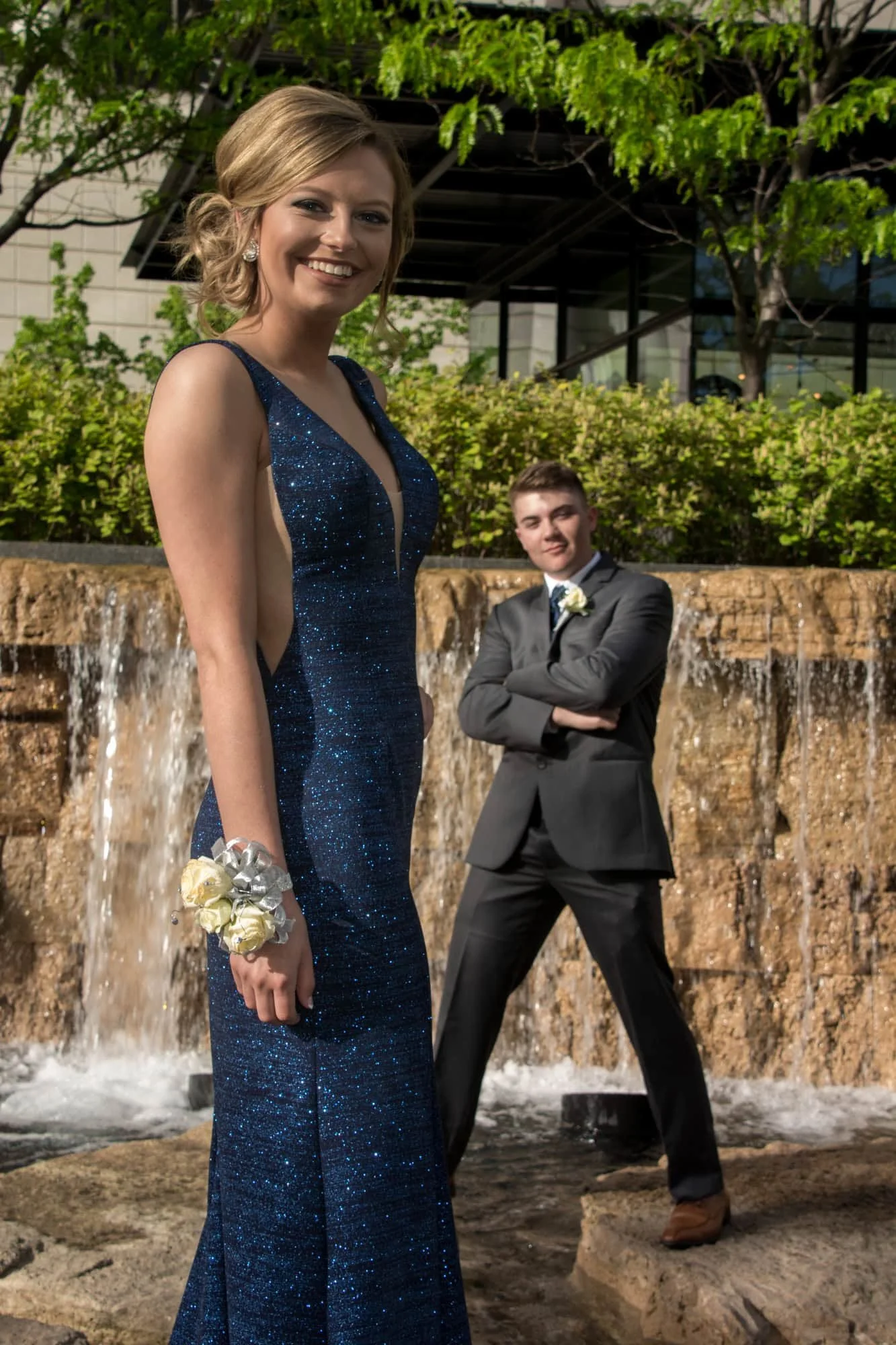 A prom girl in a sparkling blue gown posing in front of an outdoor waterfall feature while her date stands behind her with his arms crossed at Citygarden.