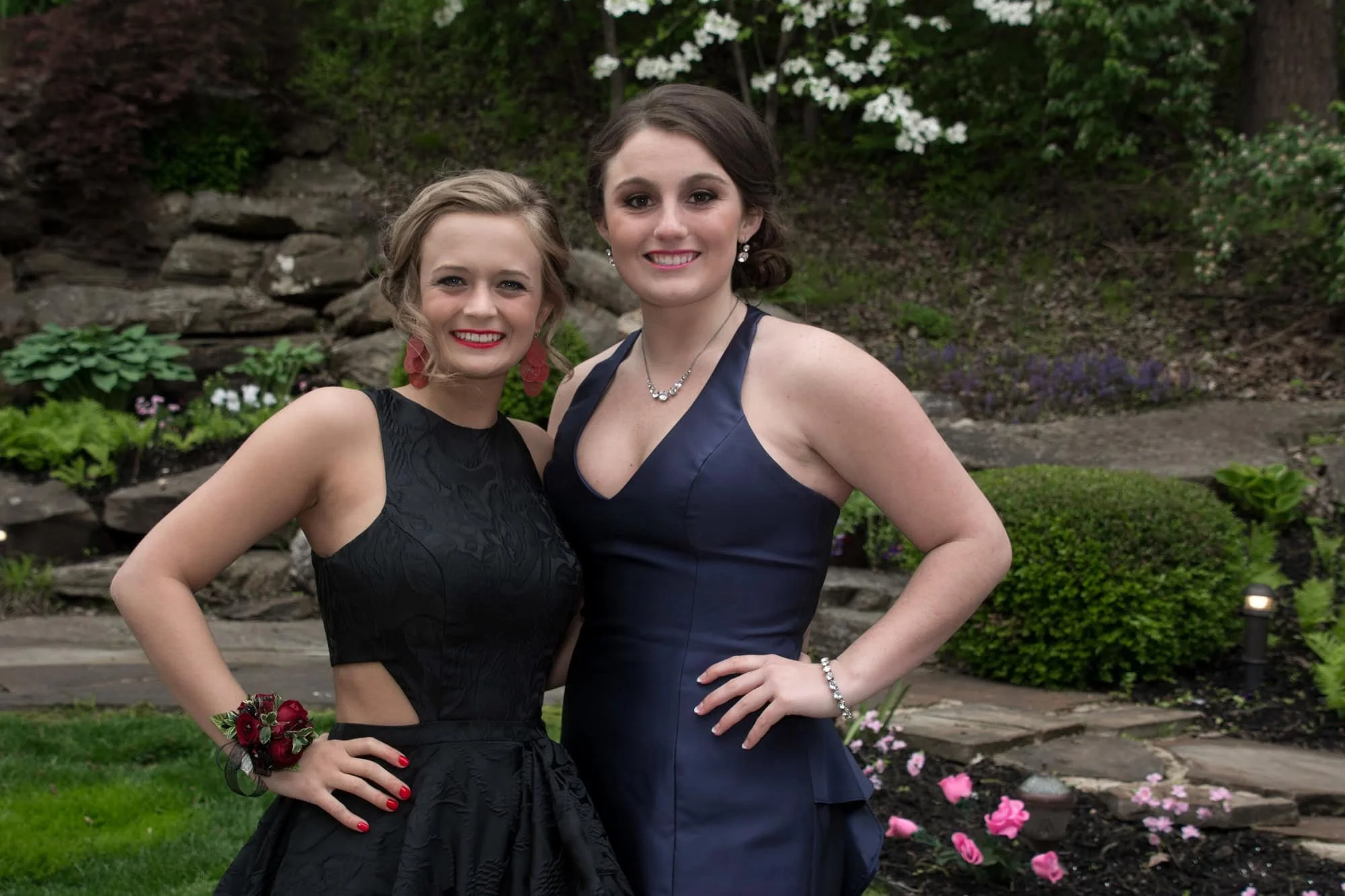 Two smiling friends posing together in formal black and navy gowns in front of a landscaped stone wall, captured by a Monroe County photographer.