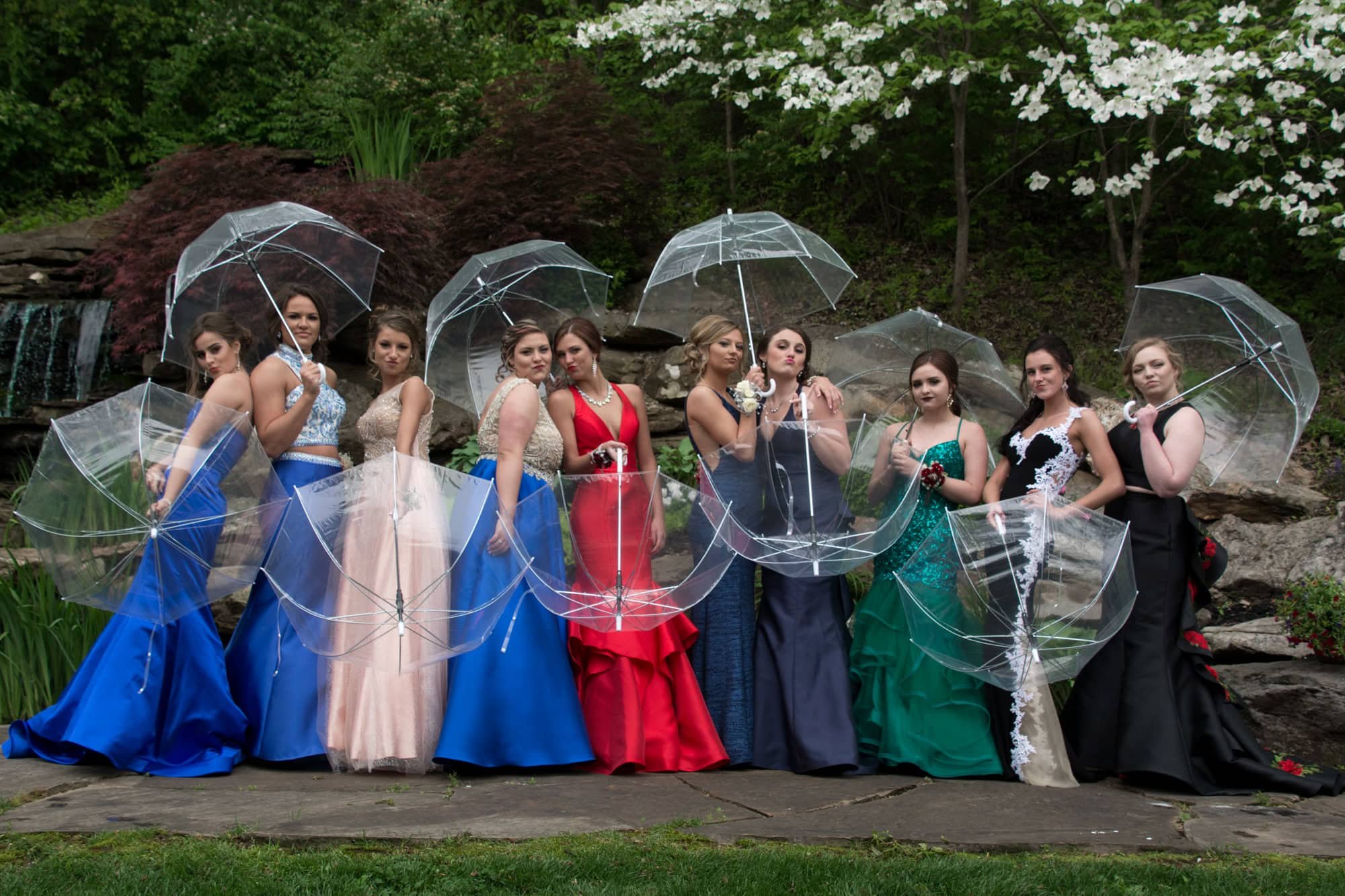 Fun group picture of prom girls in vibrant dresses smiling and holding clear umbrellas while standing outside a Monroe County prom venue on a rainy day.