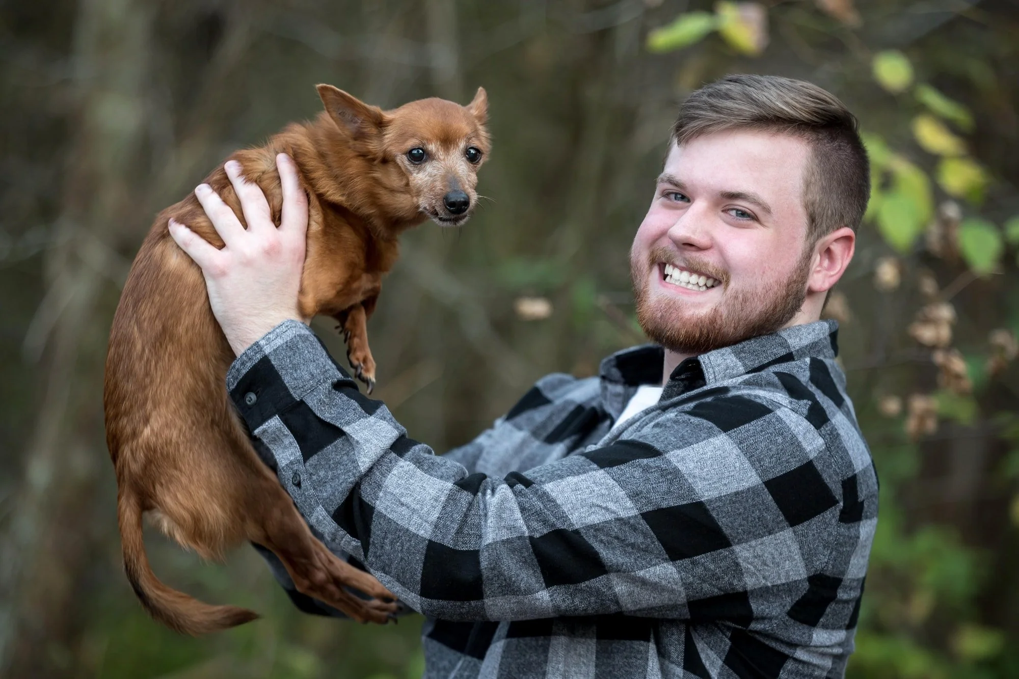 In a wooded area in Jefferson County, a high school senior guy holds his dog in the air for a fall outdoor photo.