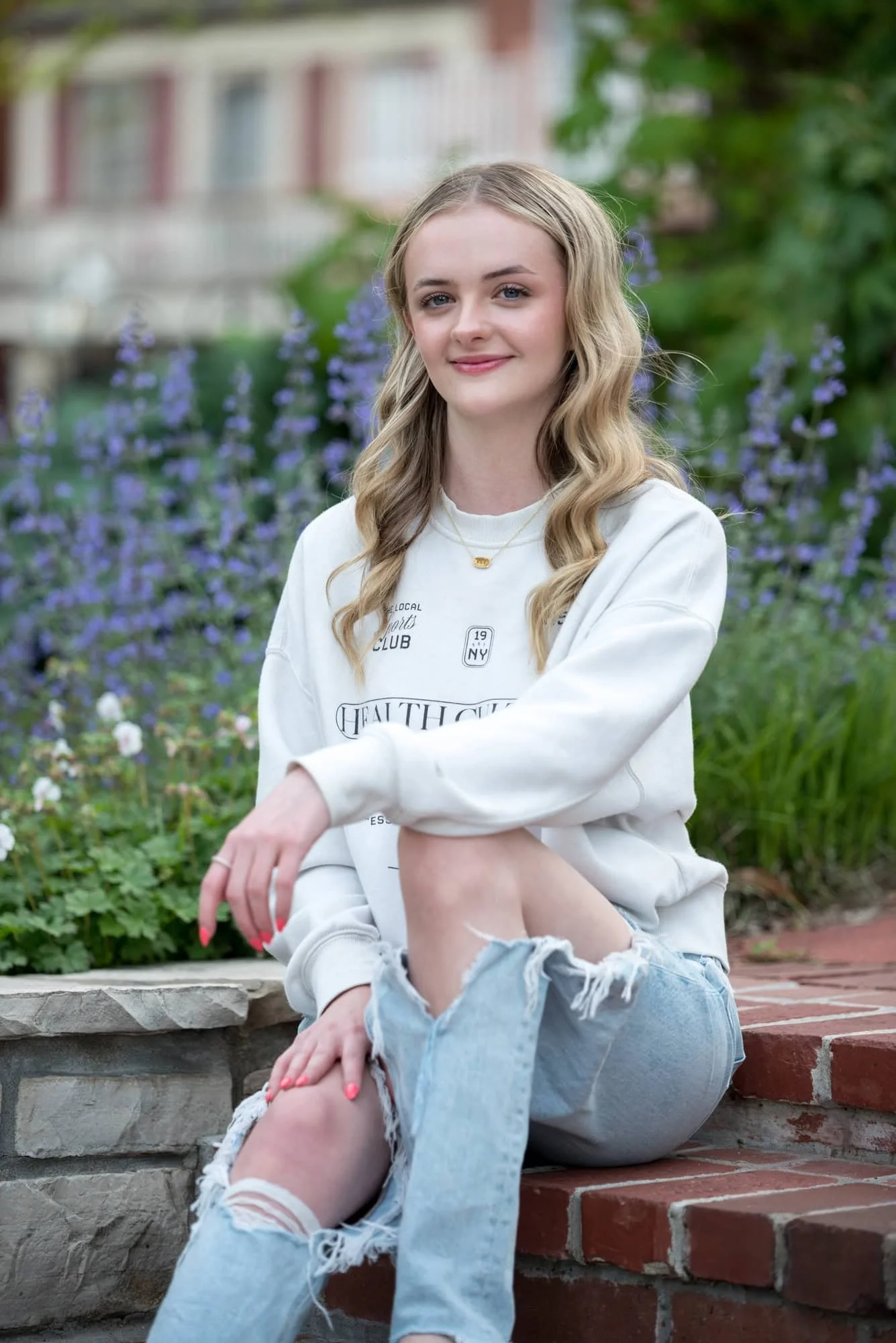 Casual and trendy senior pictures of a girl wearing a white sweatshirt and blue ripped jeans, sitting relaxed on historic brick steps in front of a blooming patch of purple flowers with her arm resting across her knee at St. Charles, MO.