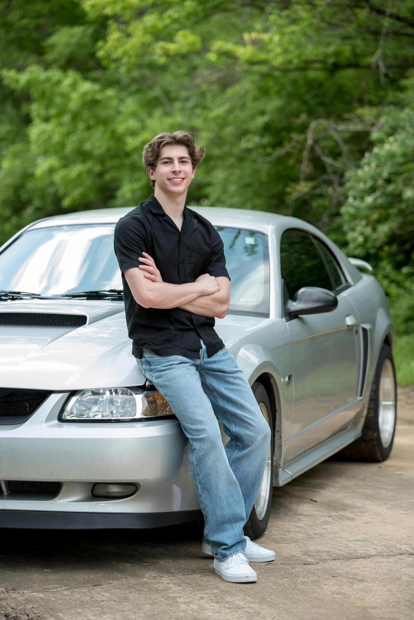Posing next to a Ford Mustang on a road, a senior guy in a black shirt and blue jeans smiles for a picture in Monroe County.