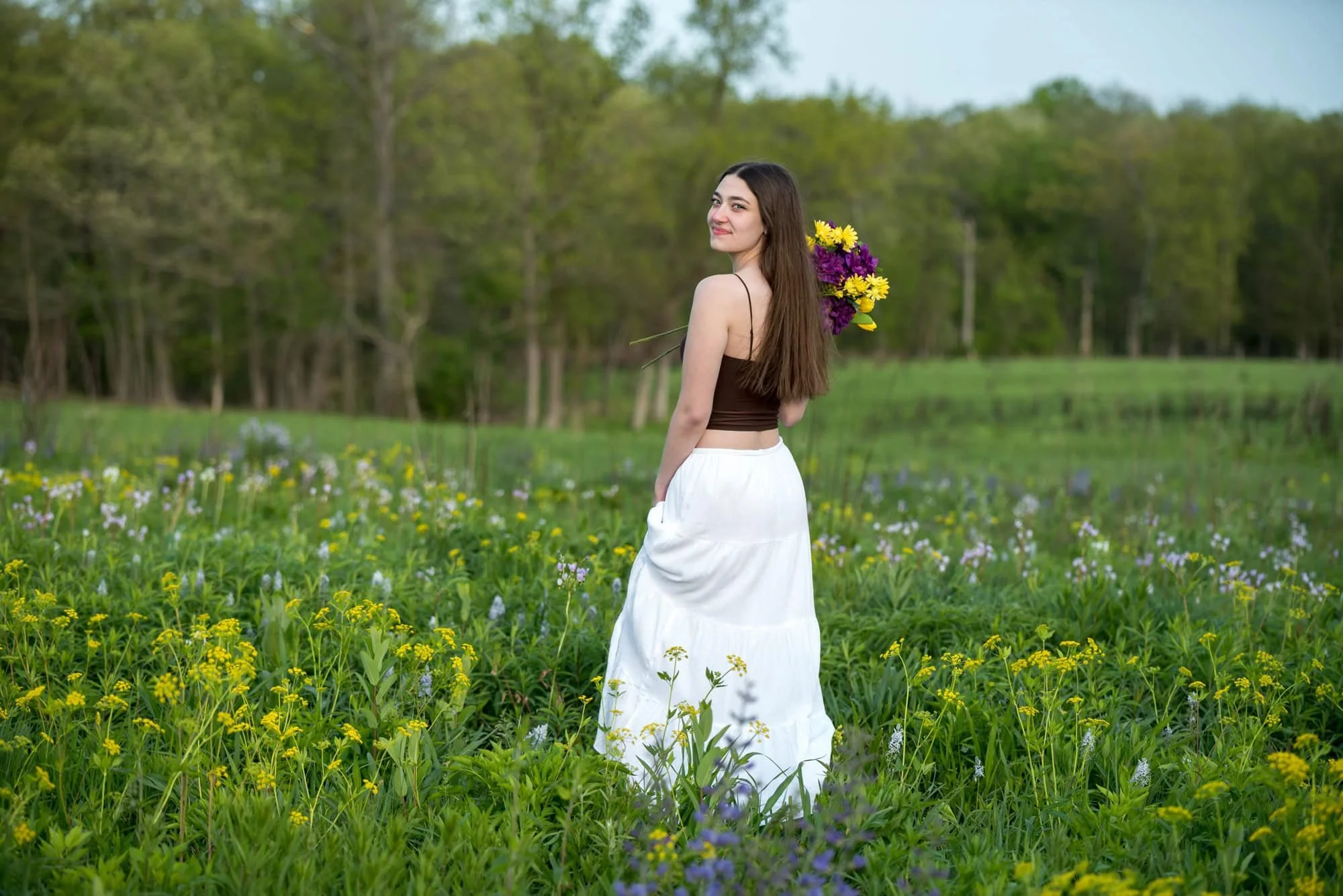 High school senior girl in a brown tank top and white skirt, holding a matching purple and yellow wildflower bouquet while looking back over her shoulder in a blooming summer field at Shaw Nature Reserve.