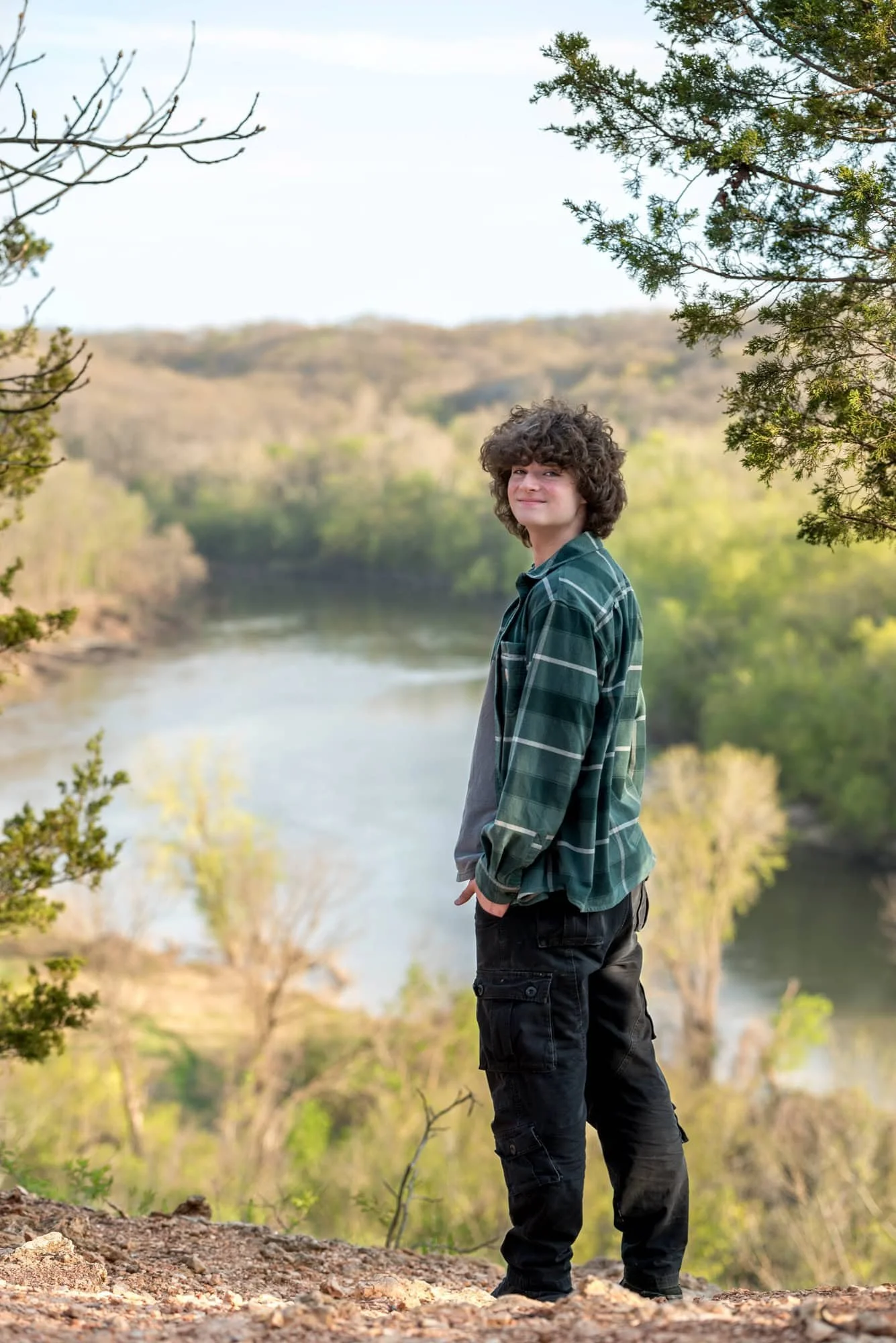 Senior guy looking back over his shoulder from the edge of a gravel overlook bluff, showcasing a sweeping view of the river and sunlit trees at Castlewood State Park in Ballwin.