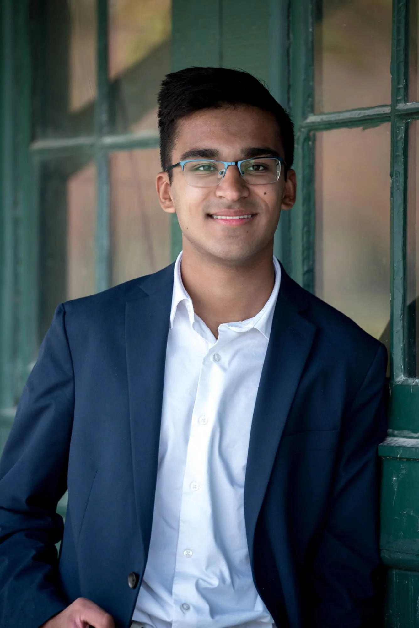 Close-up senior portrait by a West County senior photographer, showing a guy in a navy blazer leaning against a vintage green door with glass panes at Faust Park.