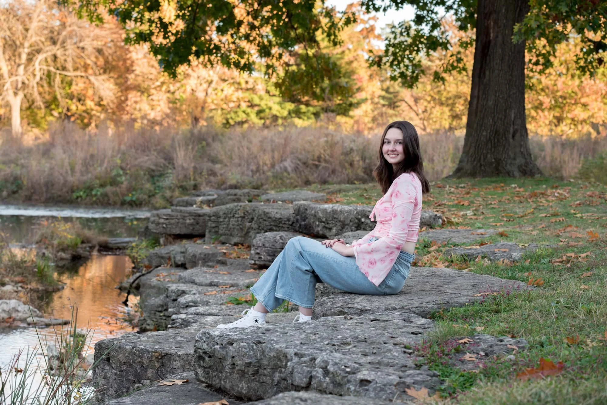 A senior girl in a pink floral crop top and blue jeans sits casually on a boulder along the water's edge for a country style picture by a St. Louis County photographer.